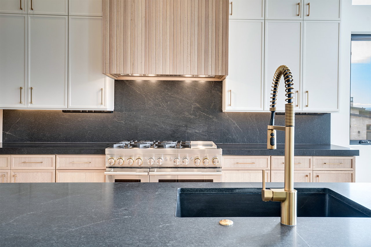 Kitchen featuring an expansive countertop with an integrated sink, a gold-toned pull-down faucet, a stainless steel range with multiple burners, a matching wood-paneled range hood, and white upper cabinetry with gold-toned hardware