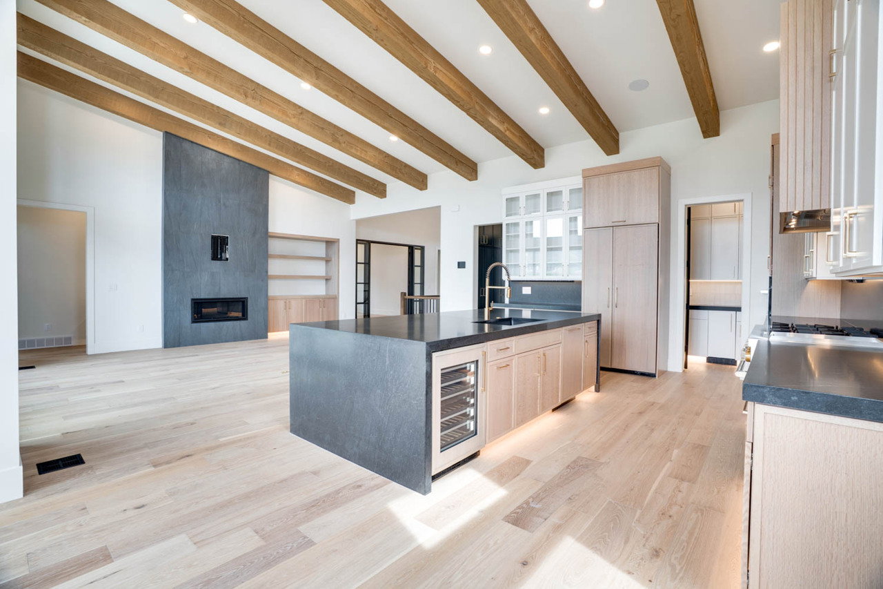 Open-concept living space featuring wood-finish flooring, exposed wood ceiling beams, a stone-clad fireplace, a central kitchen island with integrated beverage cooler, and light wood cabinetry throughout