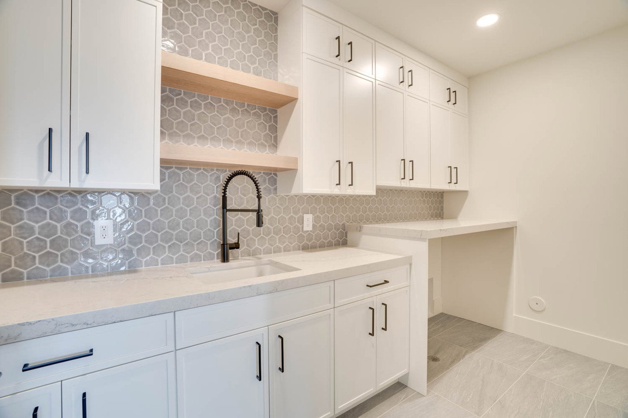 Utility room featuring an elongated sink with a matte black high-arc faucet, honeycomb tile backsplash, white shaker-style cabinetry with matte black hardware, light-toned countertops, and light gray tile flooring