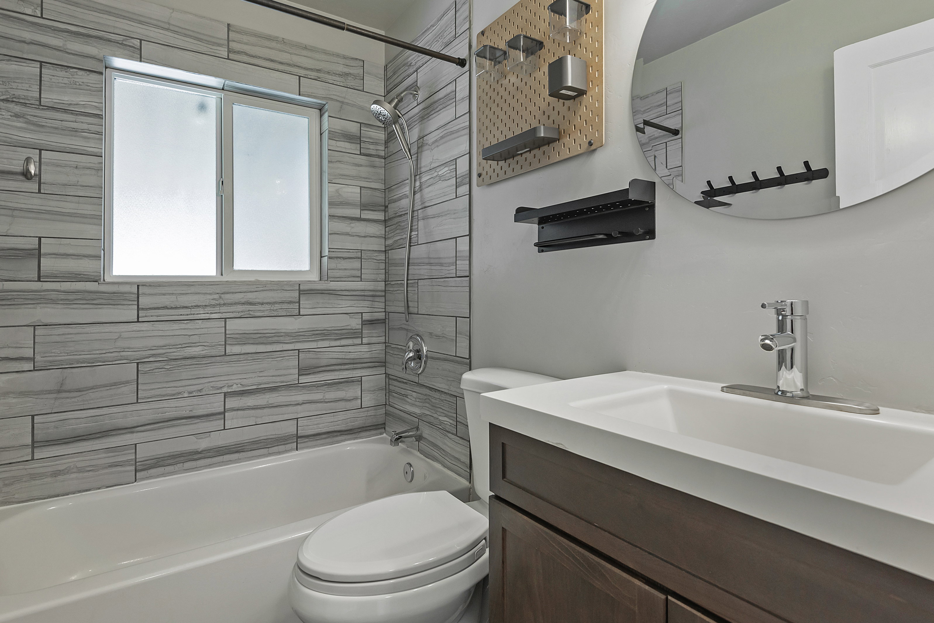 Bathroom featuring a white bathtub, tiled shower surround with a window, single vanity with integrated sink, wood-finish cabinetry, and a circular mirror