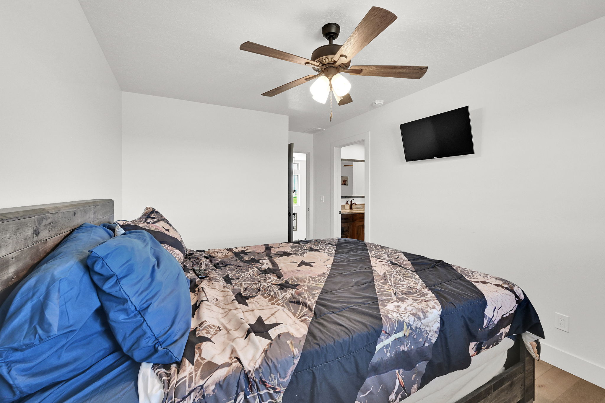 Bright bedroom featuring a ceiling fan with integrated lighting, wood-finish flooring, and a wall-mounted television