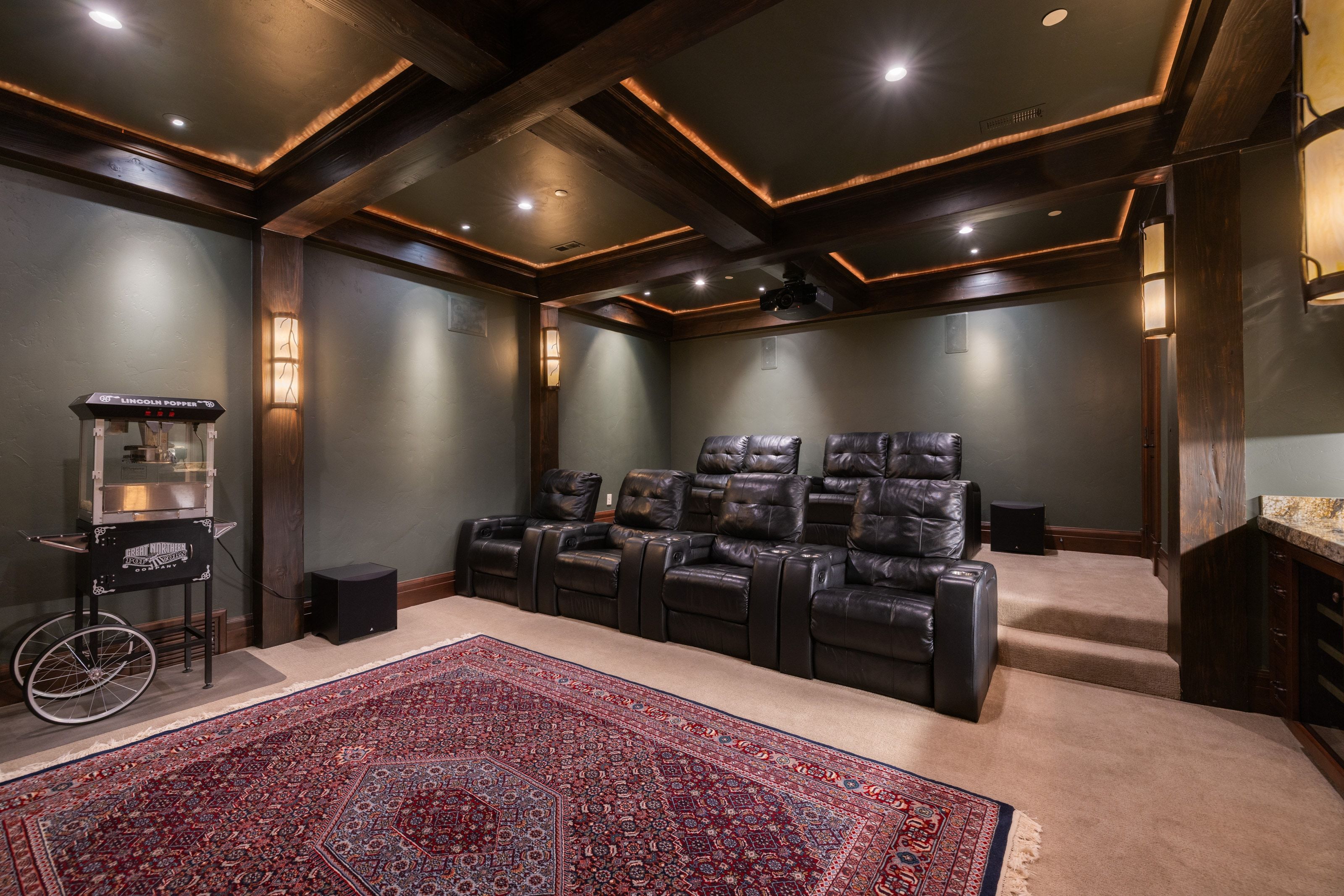 Dedicated media room featuring tiered seating, wood-beamed ceiling with recessed lighting, integrated wall sconces, and a built-in wet bar with a stone countertop