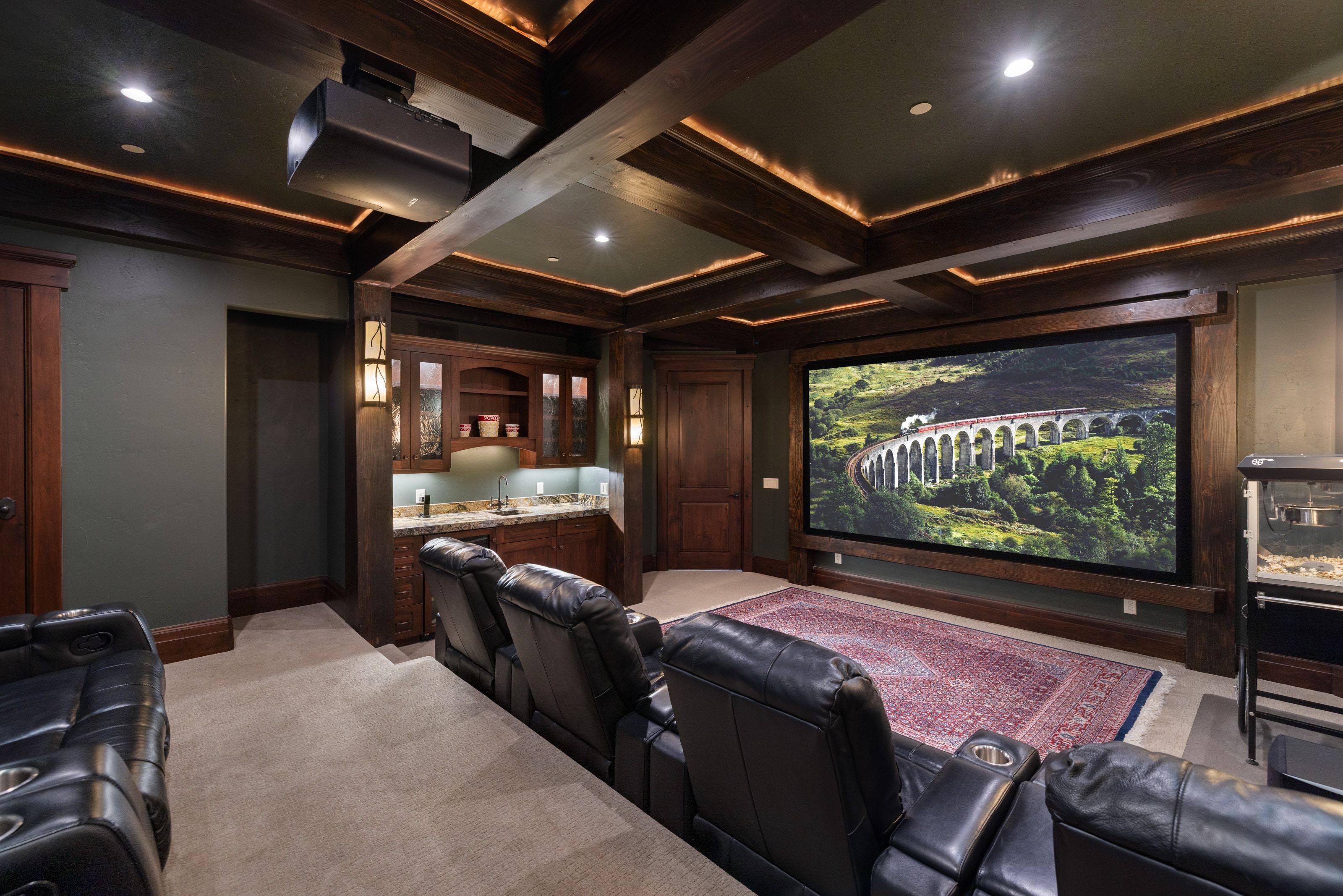 Dedicated home theater featuring a large projection screen, coffered ceiling with accent lighting, and a built-in wet bar with cabinetry
