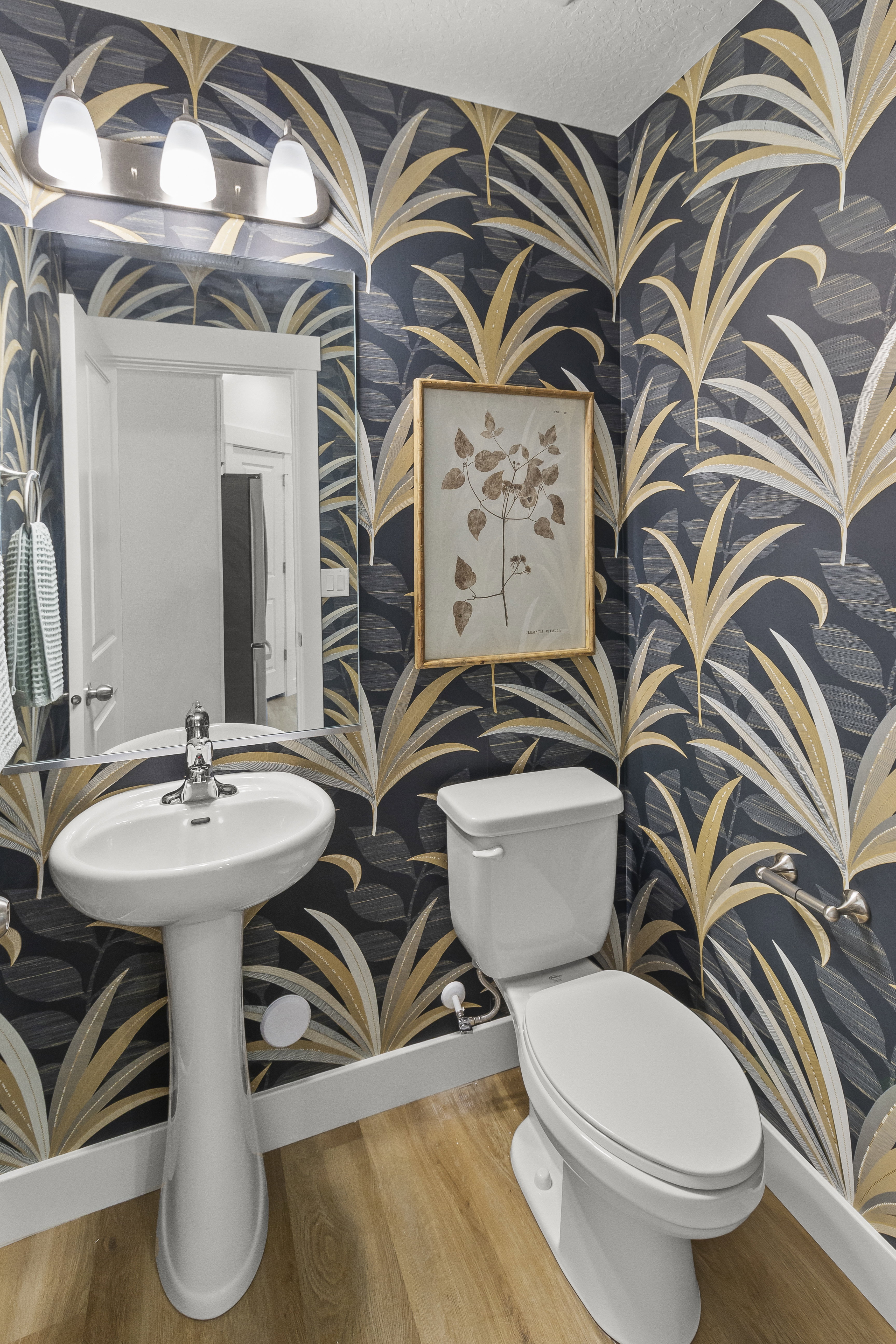 Powder room featuring botanical-patterned wallpaper, wood-finish flooring, a white pedestal sink, and a matching toilet