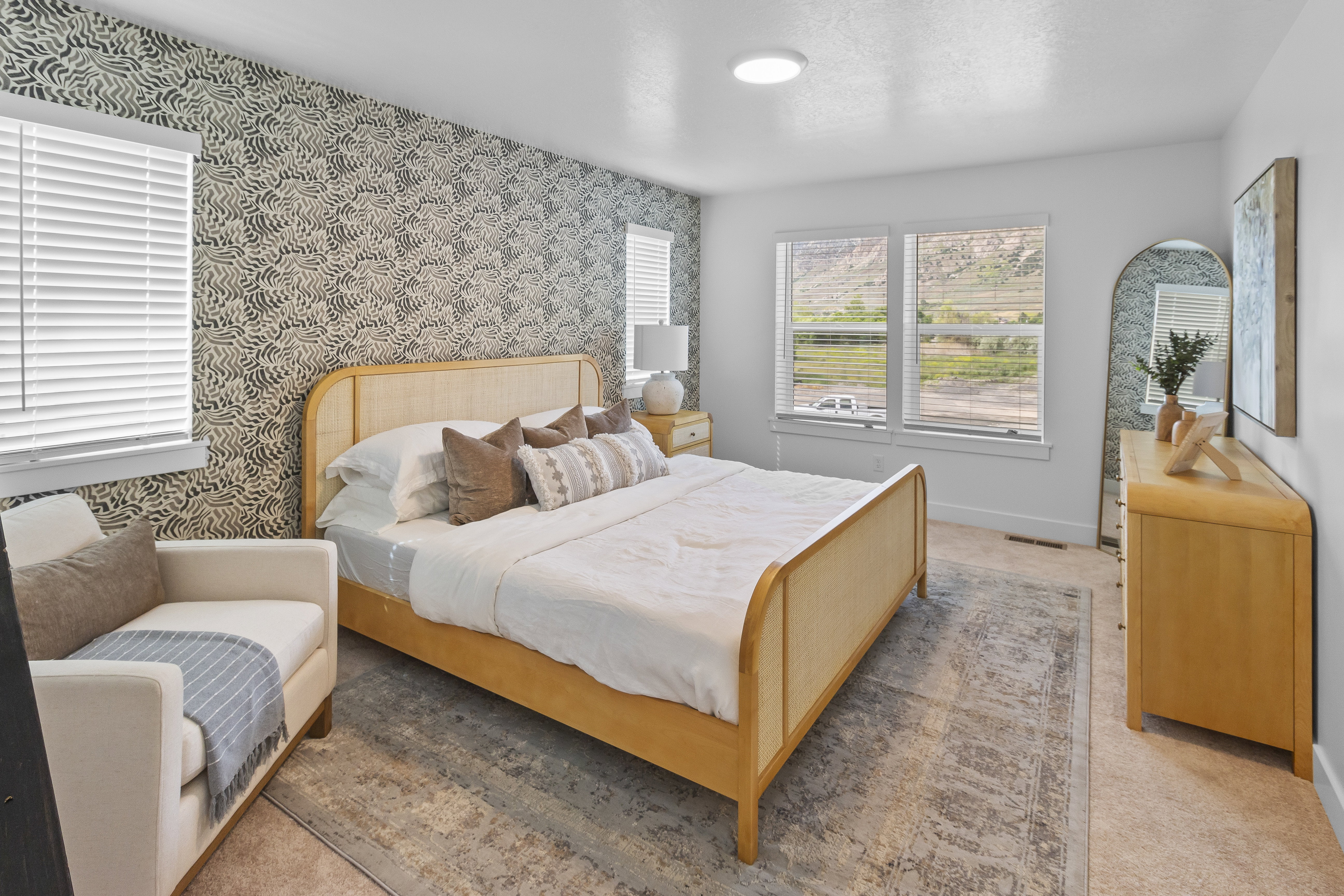 Bedroom featuring an accent wall with patterned wallpaper, two windows with blinds, and a light-colored area rug over carpeting