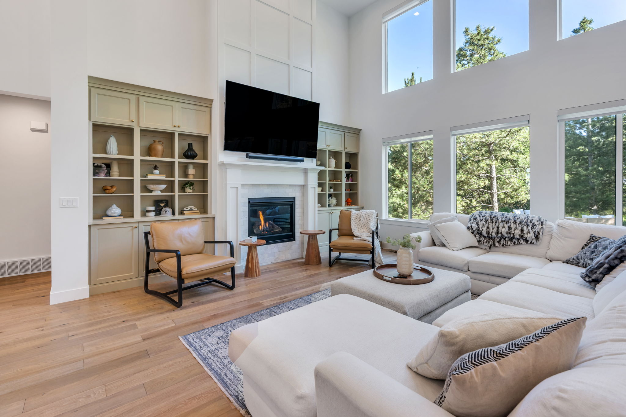 Spacious living area featuring expansive windows, a fireplace with a stone surround, integrated built-in shelving, wood-finish flooring, and a soaring ceiling with paneling accents
