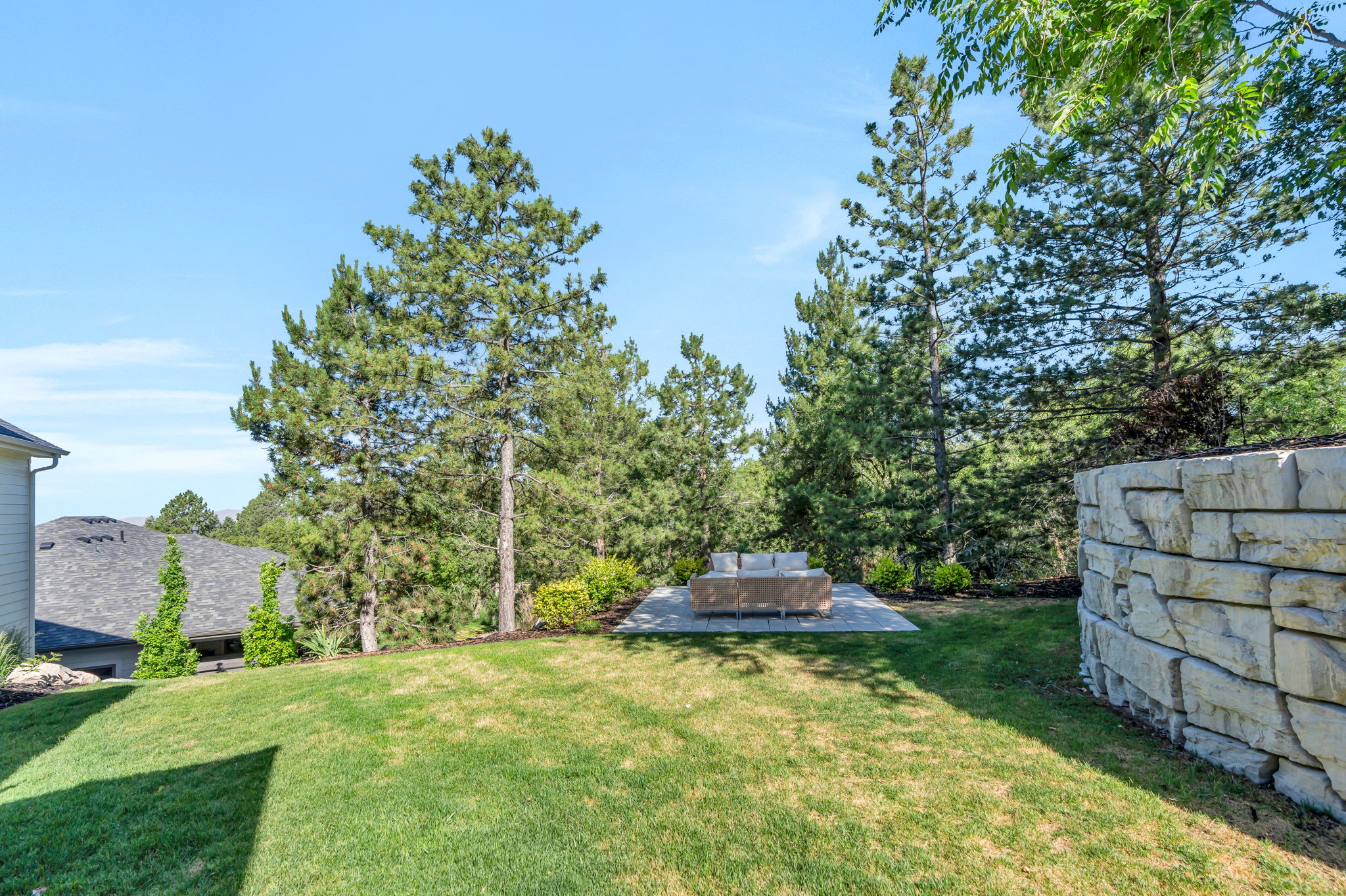 Expansive yard featuring a stone retaining wall, mature pine trees, and a paver patio