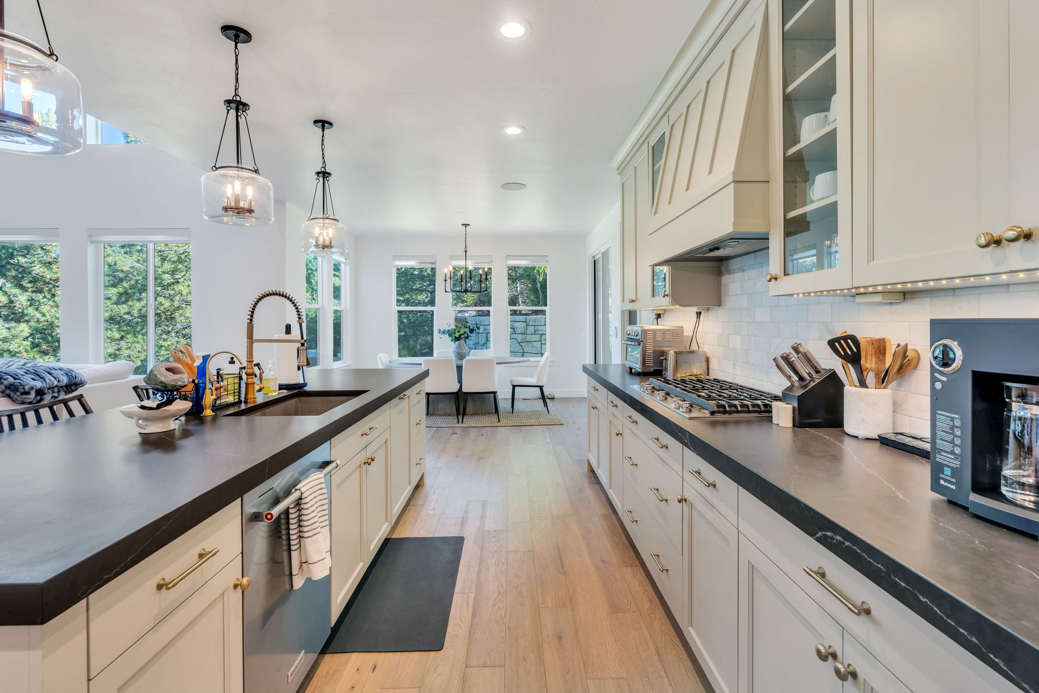 Open-concept kitchen featuring dark countertops, light cabinetry with gold-tone hardware, wood-finish flooring, a built-in gas range, and a spacious island with an integrated sink