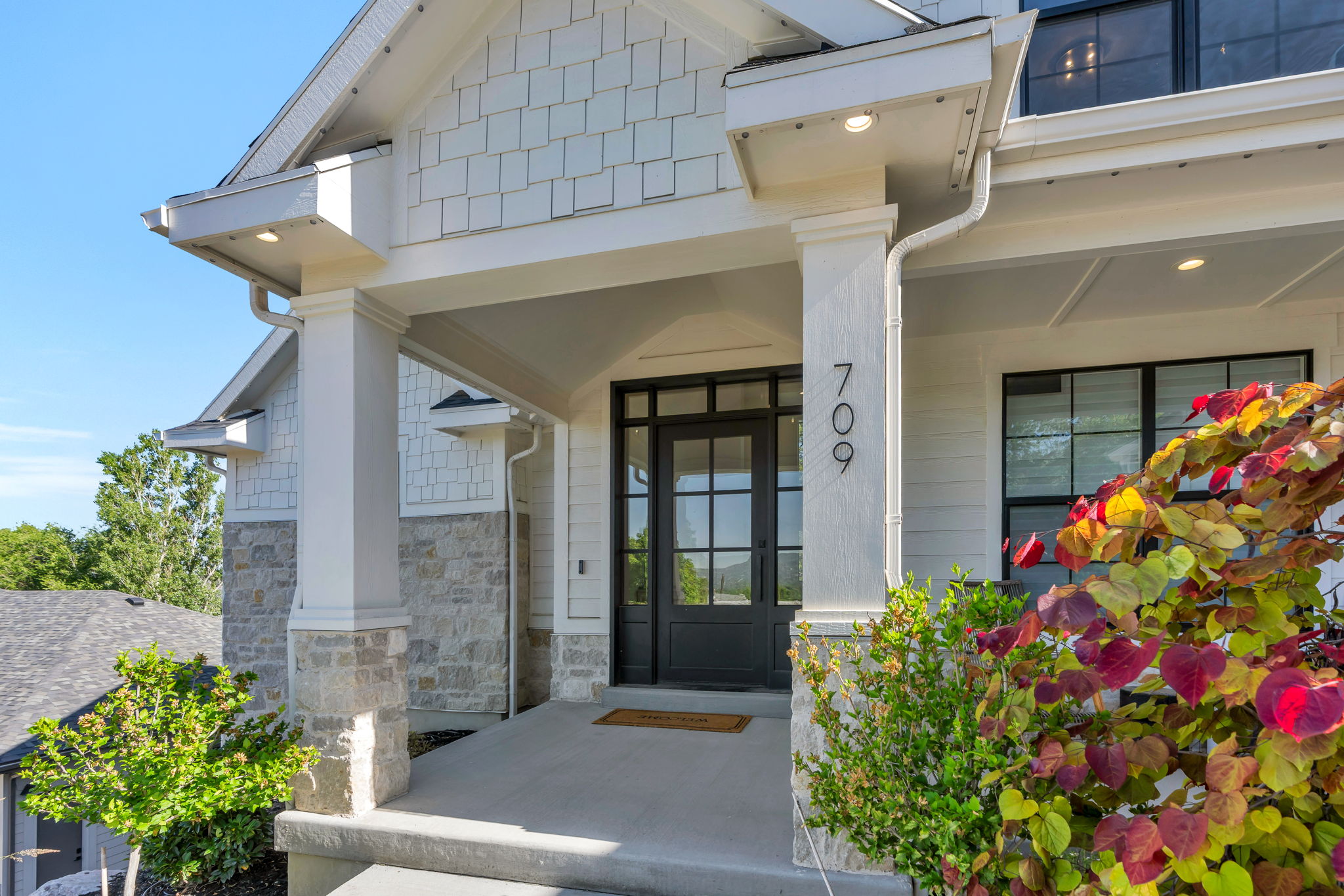 Covered front porch featuring a dark-framed glass paneled entry door, stone veneer, shingle siding, and integrated soffit lighting