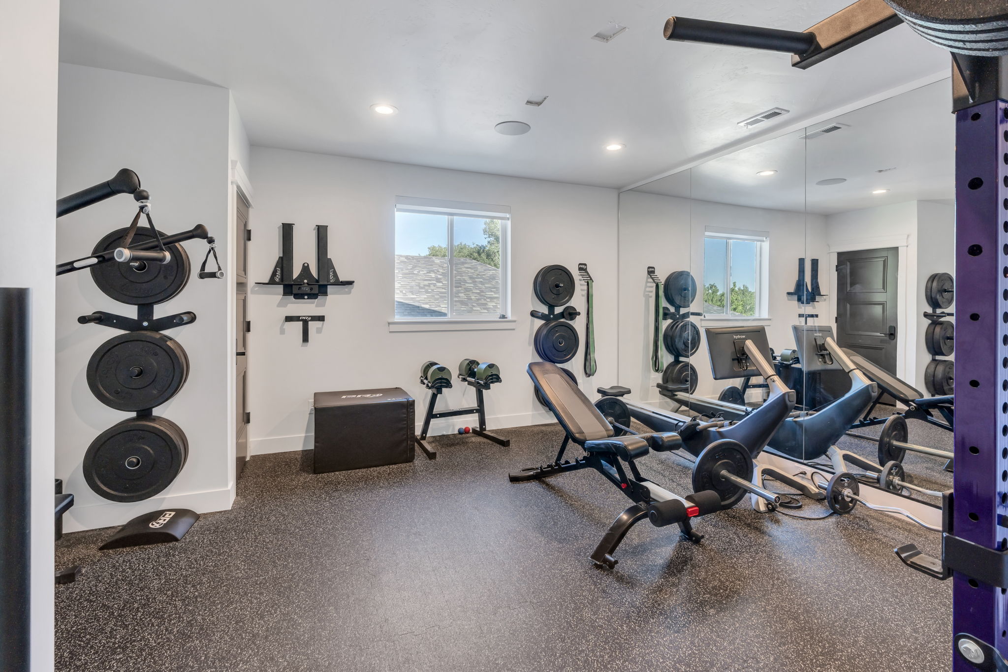 Dedicated fitness space featuring impact-absorbing rubber flooring, a large wall-mounted mirror, multiple windows, and recessed lighting
