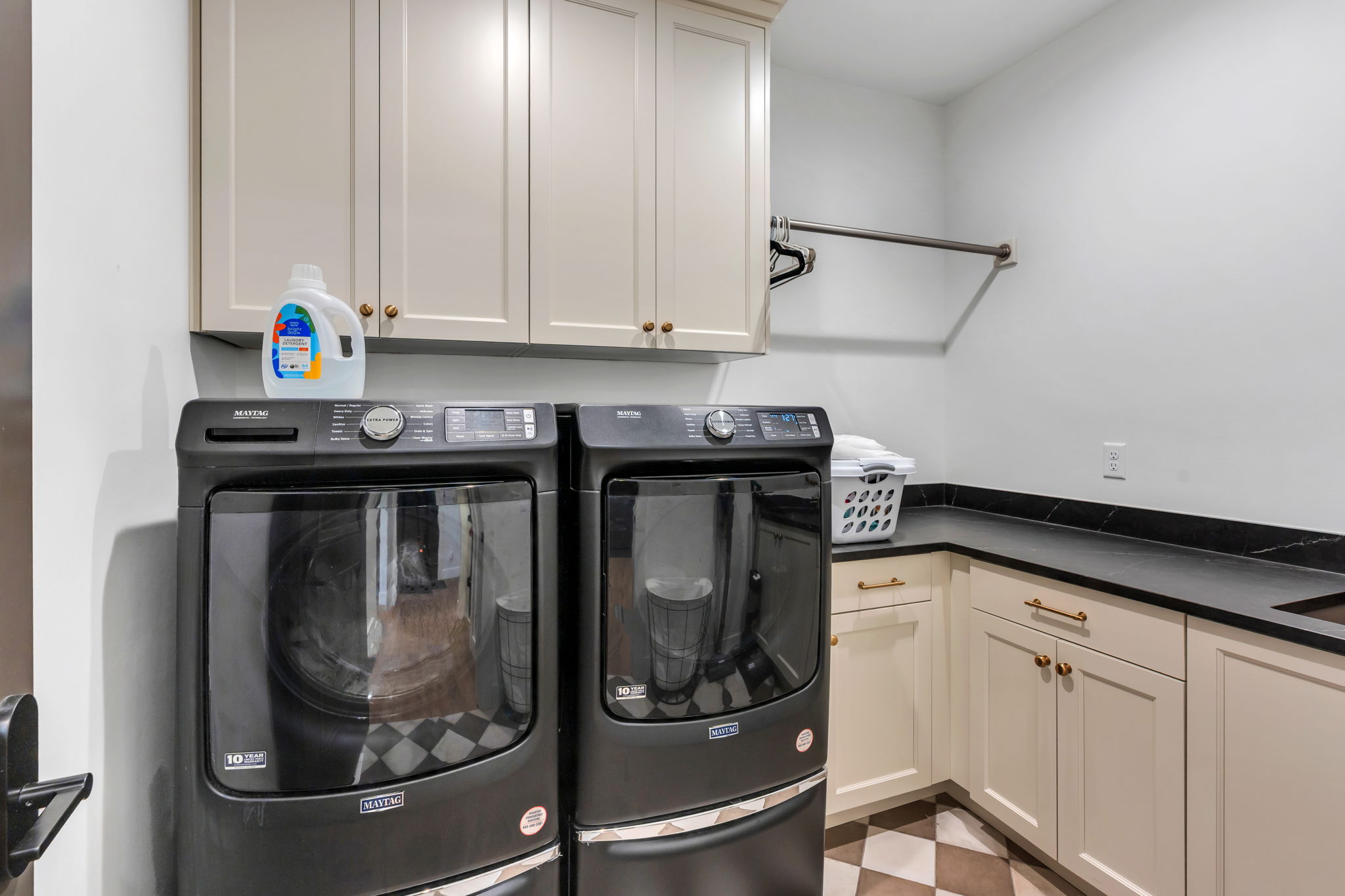 Laundry area featuring a checkered tile floor, light-colored cabinetry with brass hardware, dark countertops, upper wall cabinets, and a built-in hanging rod