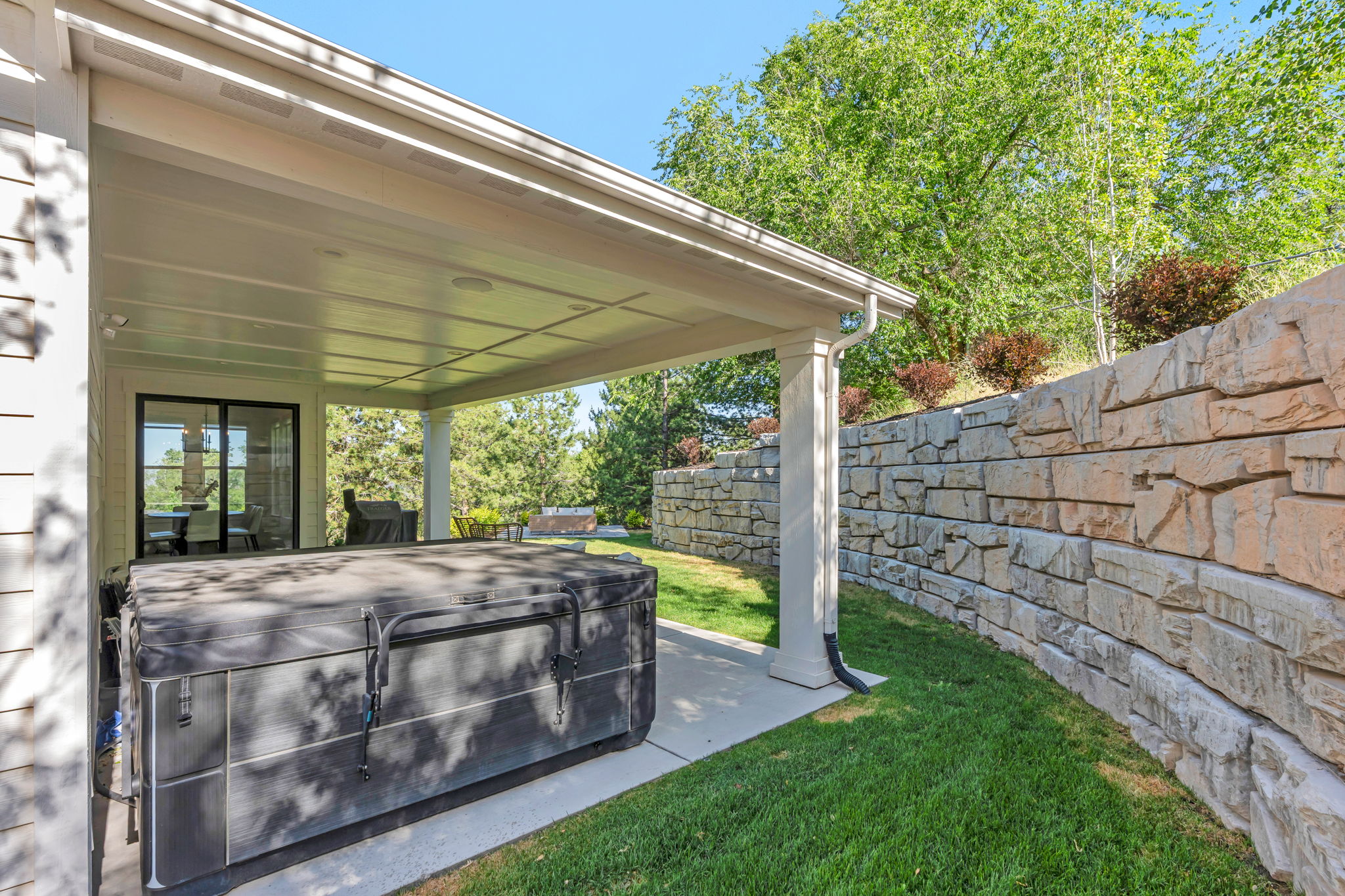 Covered patio area featuring a stone retaining wall, mature trees, and a grassy expanse
