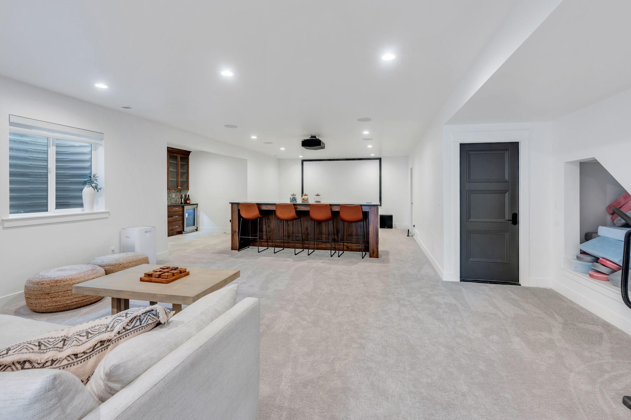 Spacious entertainment area featuring light-toned carpeting, a built-in wet bar with a wood-finish countertop, a dedicated projection screen, recessed lighting, and a window with horizontal blinds