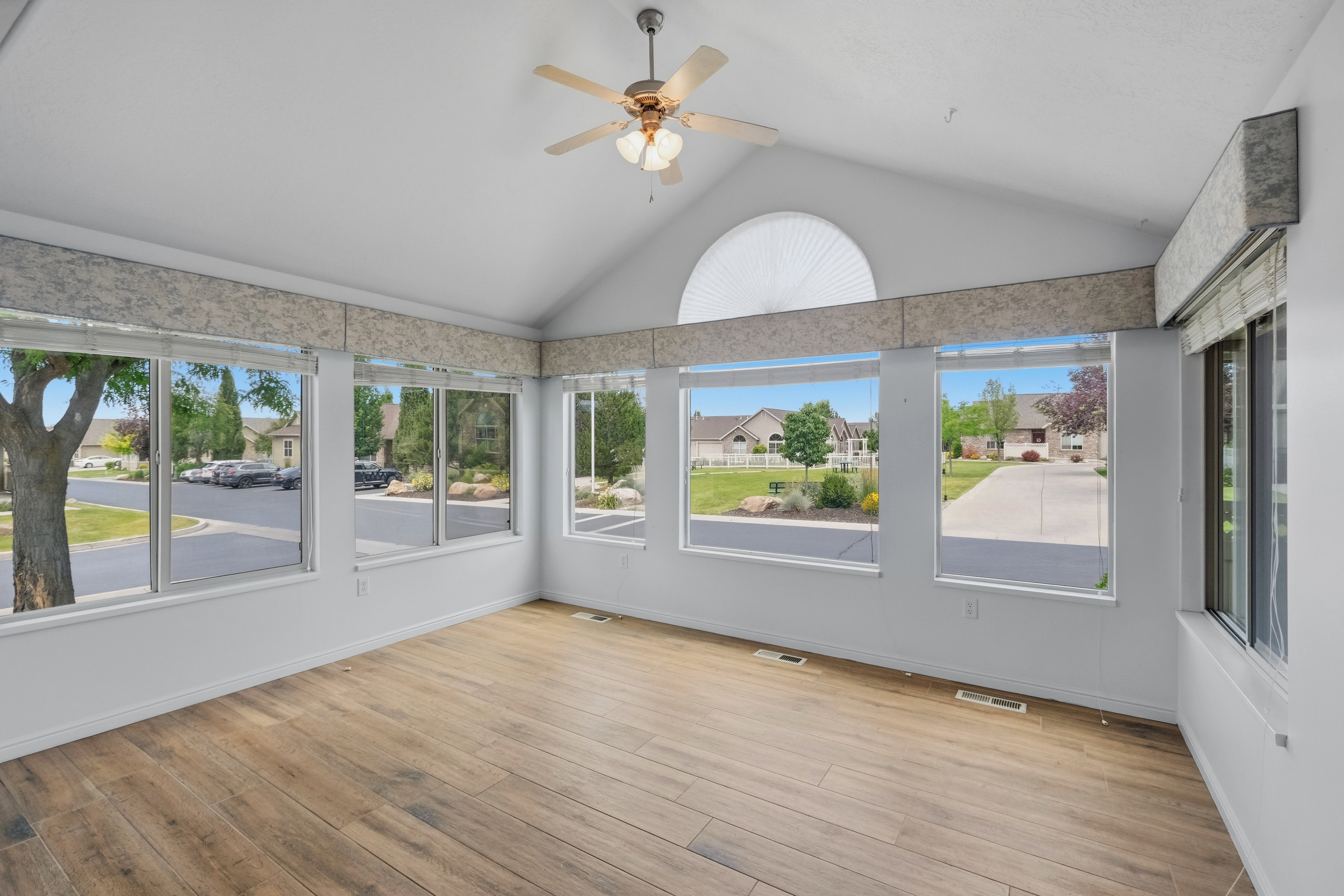Spacious room featuring vaulted ceilings, wood-finish flooring, a ceiling fan with integrated lighting, and multiple windows with valances