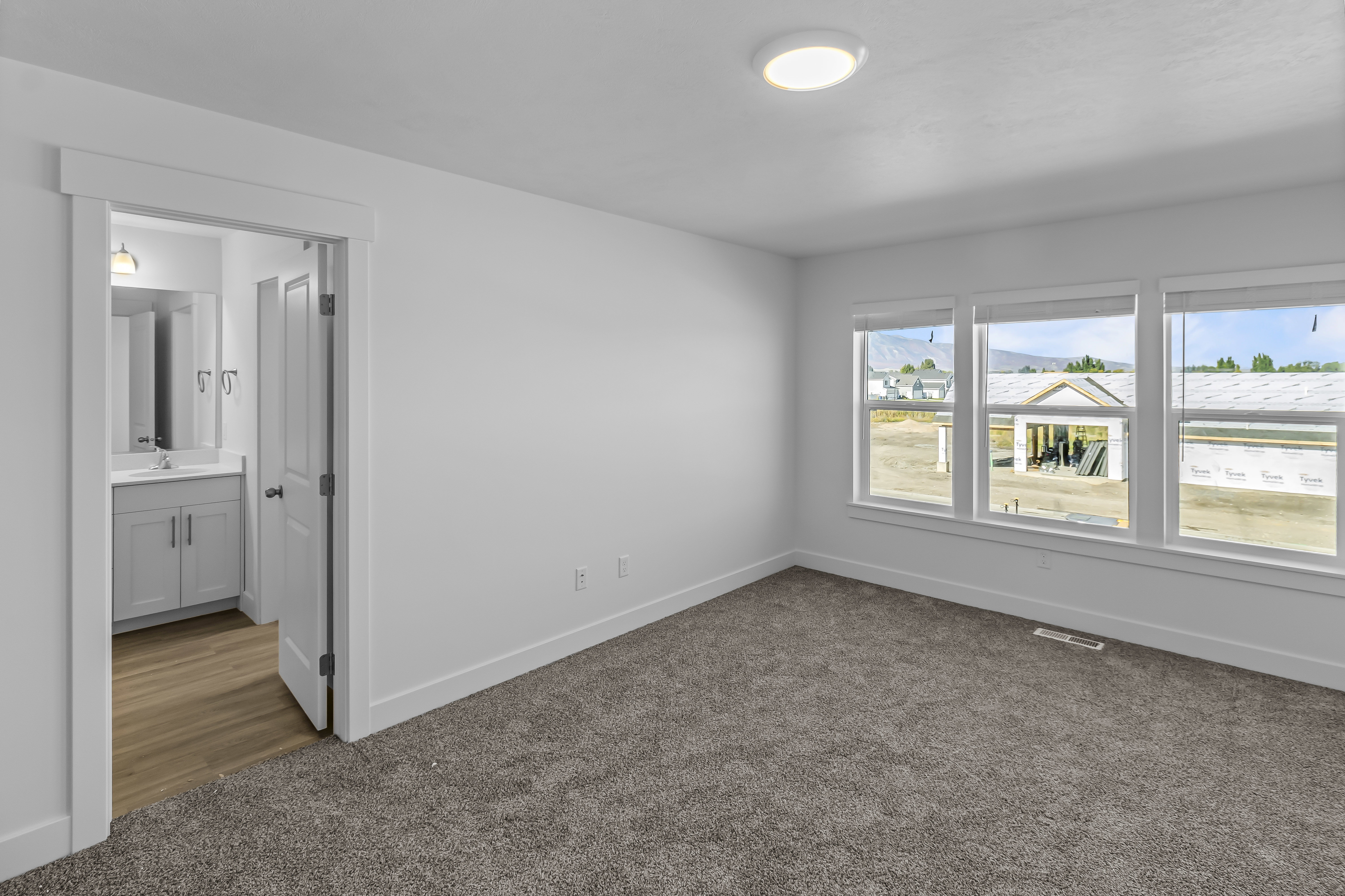 Spacious room with a neutral color palette, grey carpet flooring, three large windows, and an en-suite bathroom featuring a single vanity with a light grey cabinet