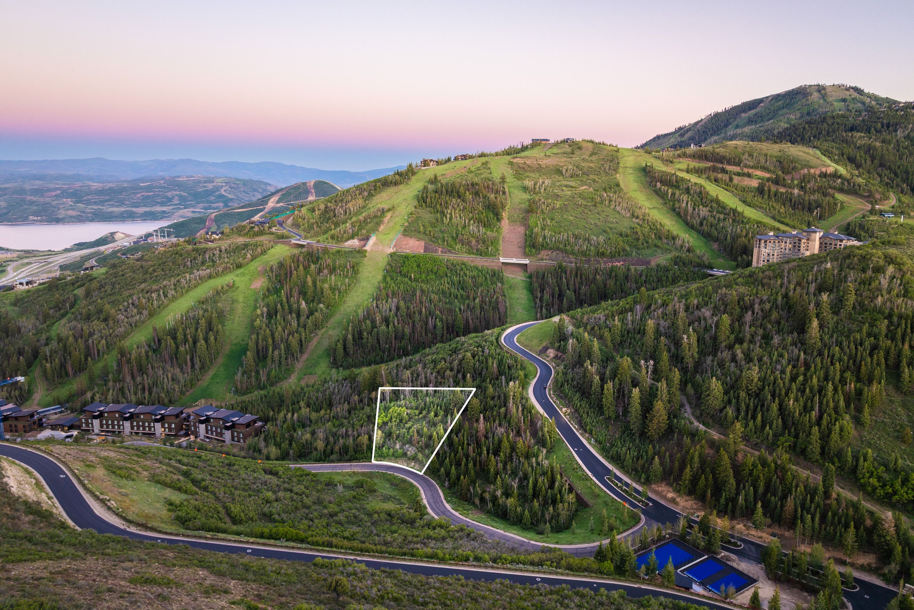 Wooded mountain lot featuring a curved paved road, surrounding evergreen trees, and distant water