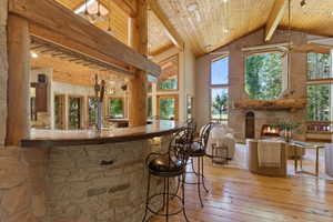 Great room featuring a prominent stone fireplace with a log mantle, soaring vaulted wood-paneled ceilings, expansive windows, wood-finish flooring, and a built-in stone-faced bar