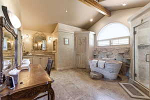 Stone soaking tub with rock surround, glass-enclosed shower, arched window with plantation shutters, vaulted ceiling with exposed wood beam, and dual vanity with ornate mirrors