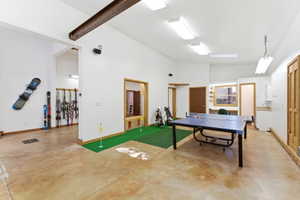 Spacious recreation area featuring concrete flooring, a built-in sink with cabinetry, a dedicated work area with a window, and a high vaulted ceiling with exposed beam