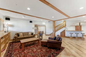 Spacious living area featuring light wood-finish flooring, exposed wood ceiling beams, a built-in wet bar, and a prominent wood staircase