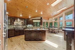Open-concept kitchen and dining area featuring a vaulted wood-paneled ceiling, wood-finish flooring, a central island with an integrated sink, stainless steel appliances, and a wall of floor-to-ceiling windows
