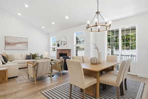 Open-concept living and dining area featuring wood-finish flooring, a vaulted ceiling with recessed lighting, and a brick-faced fireplace with a white mantel