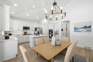 Expansive kitchen and dining area featuring light wood-finish flooring, white cabinetry, a kitchen island with seating, stainless steel appliances, and a prominent circular chandelier