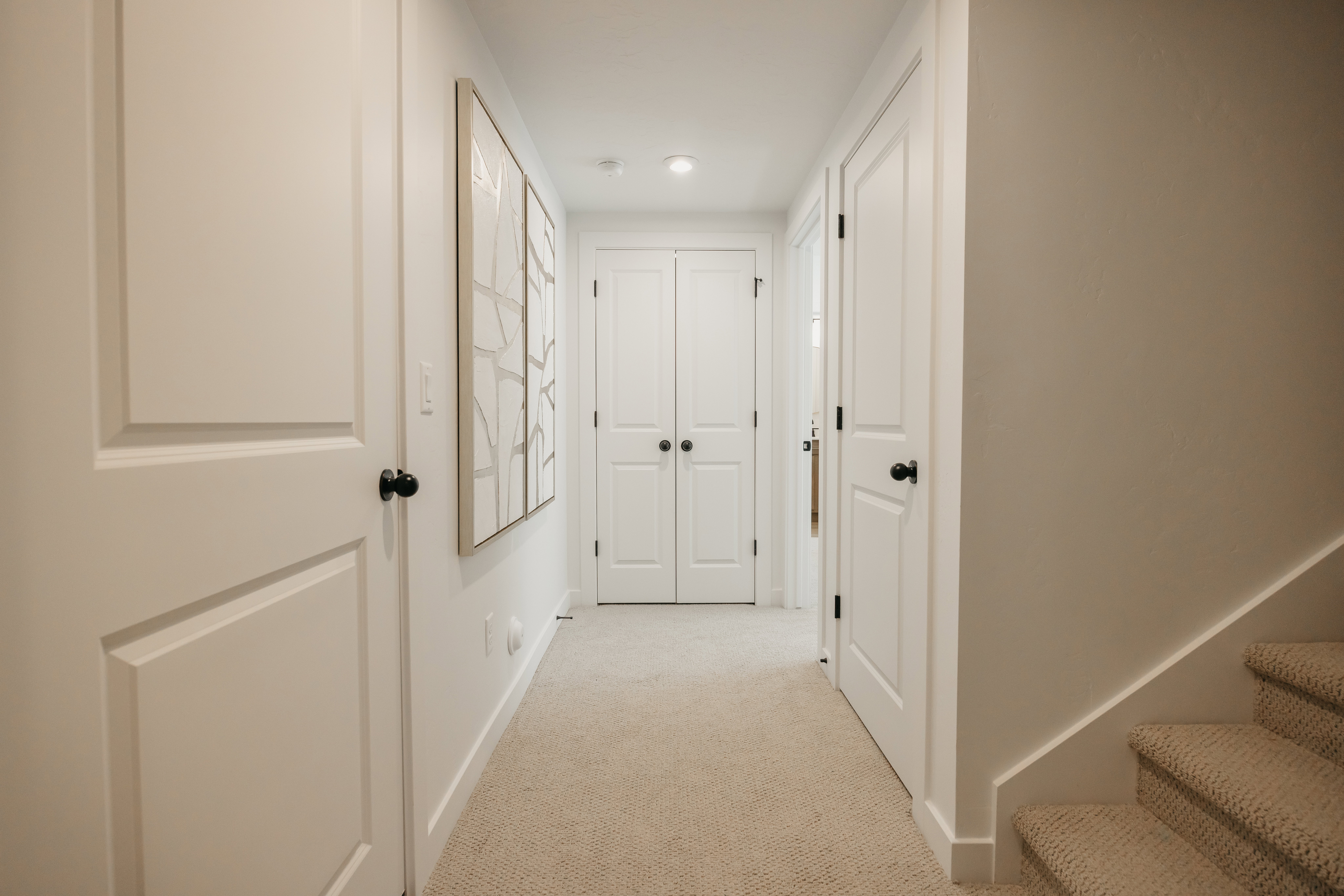 Carpeted hallway with flush-mounted lighting, multiple paneled doors with matte black hardware, and a staircase featuring carpeted treads and risers