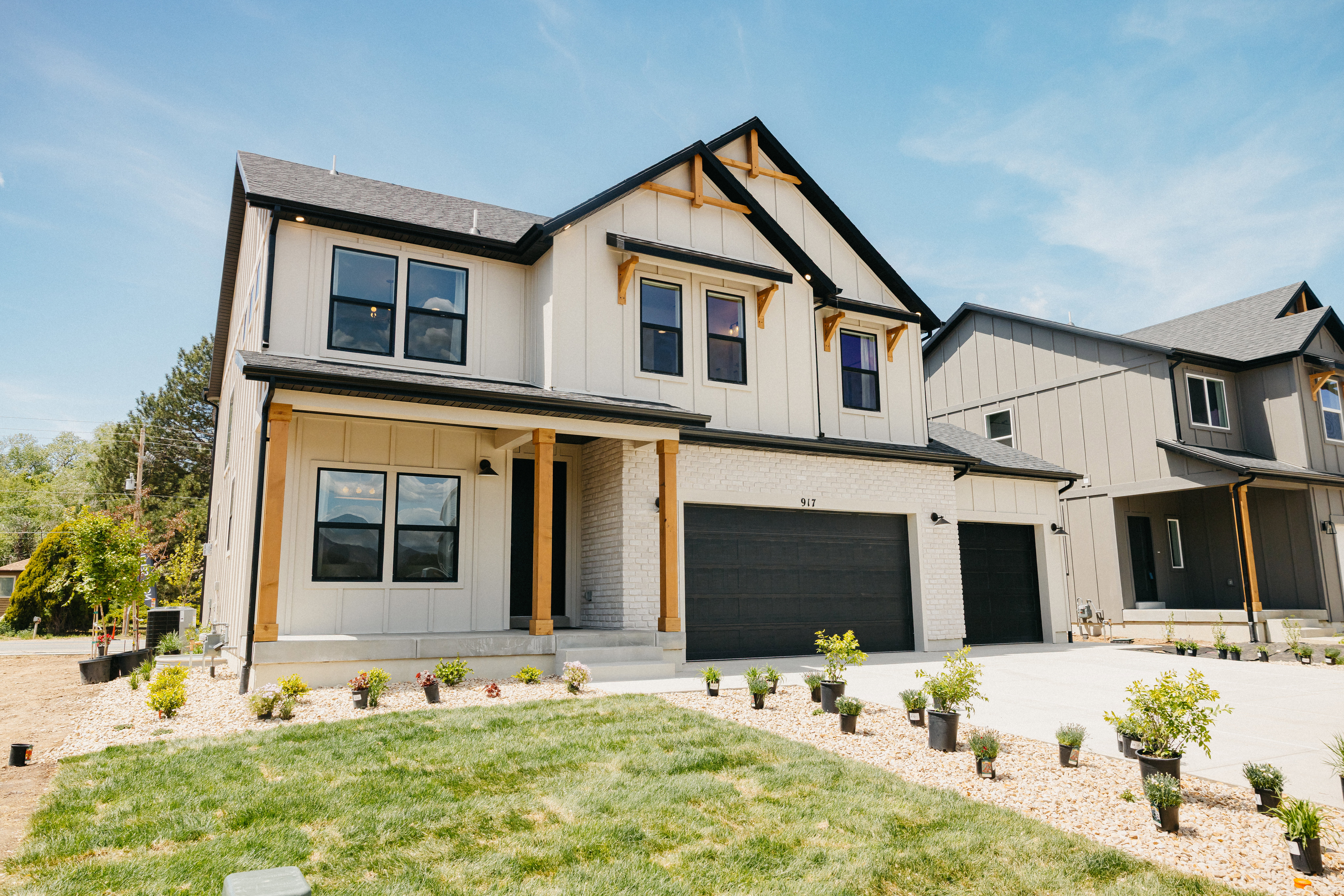 Modern farmhouse exterior featuring light-colored siding with dark trim, a two-car garage, an additional single-car garage, and an inviting covered porch supported by exposed wood beams