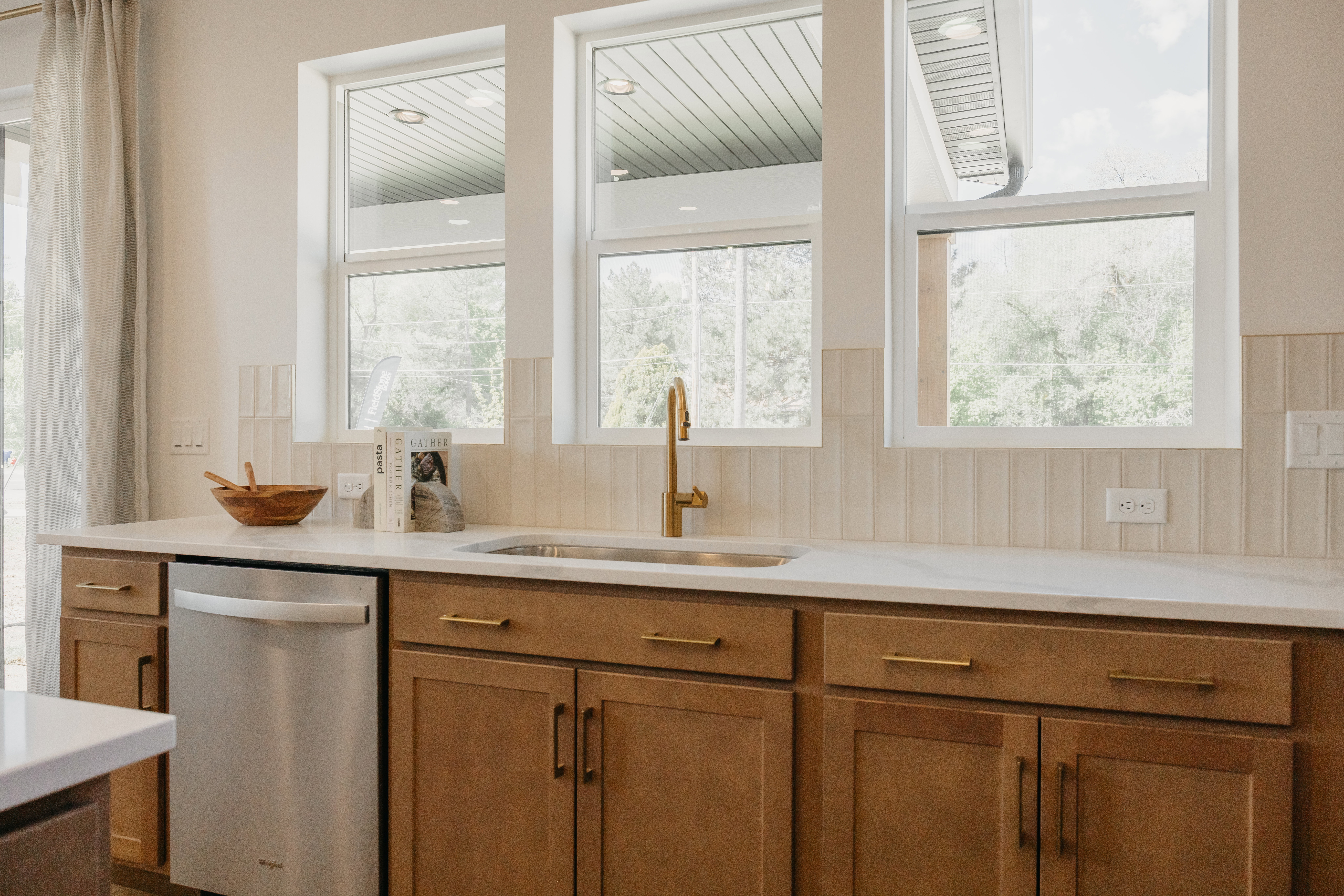 Kitchen sink area featuring three large windows, a gold-toned faucet, light wood-finish cabinetry, and a stainless steel dishwasher