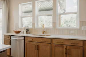 Kitchen sink area featuring three large windows, a gold-toned faucet, light wood-finish cabinetry, and a stainless steel dishwasher