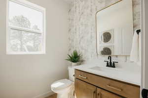 Contemporary bathroom featuring a single window, a toilet, a vanity with a white countertop and integrated sink, a rectangular gold-framed mirror, and patterned accent wallpaper