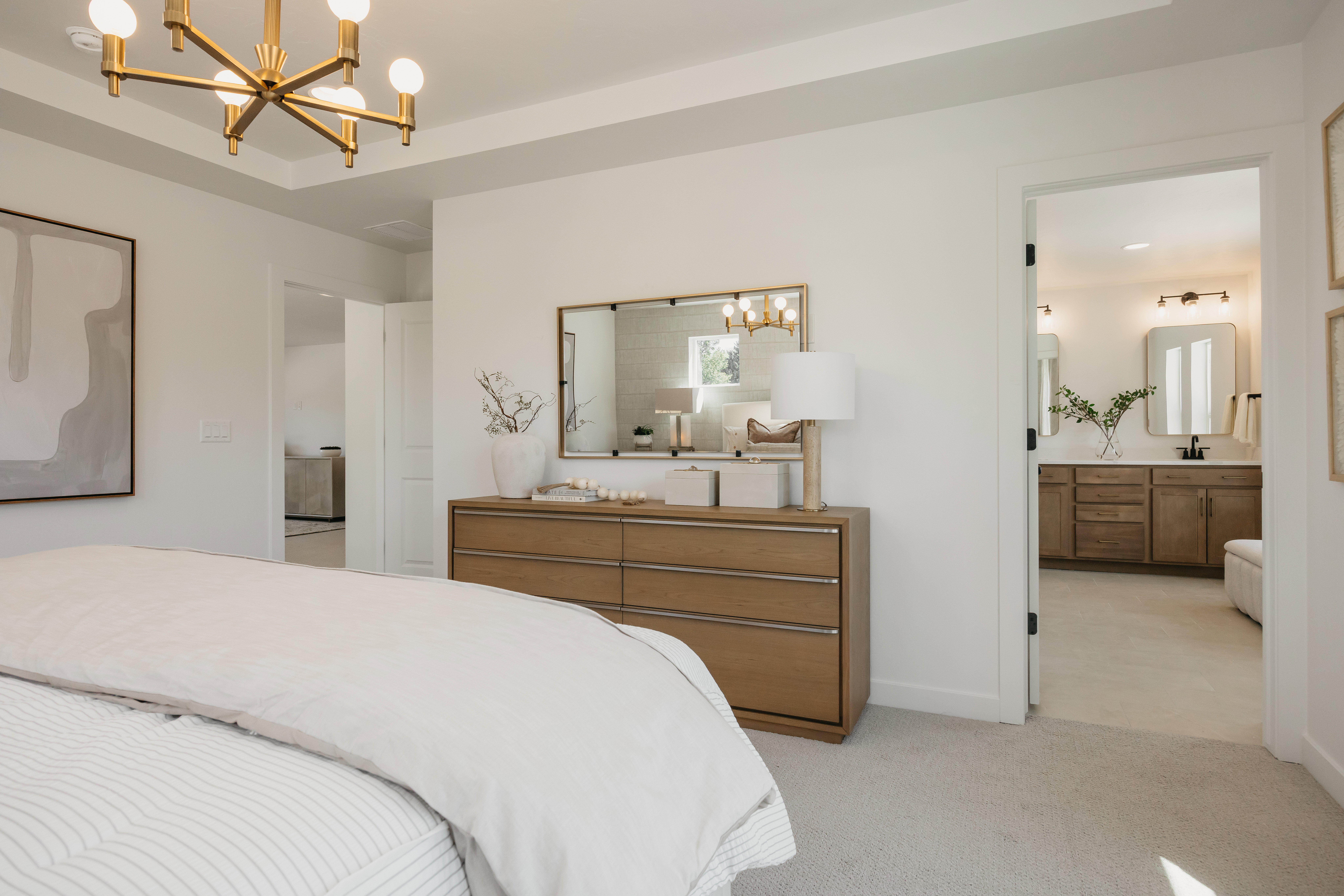 Tray ceiling with a modern gold-finish chandelier, soft gray carpet, and a light wood-finish dresser with a gold-framed mirror