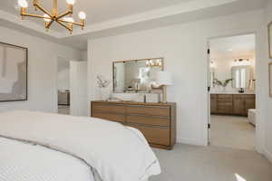 Tray ceiling with a modern gold-finish chandelier, soft gray carpet, and a light wood-finish dresser with a gold-framed mirror