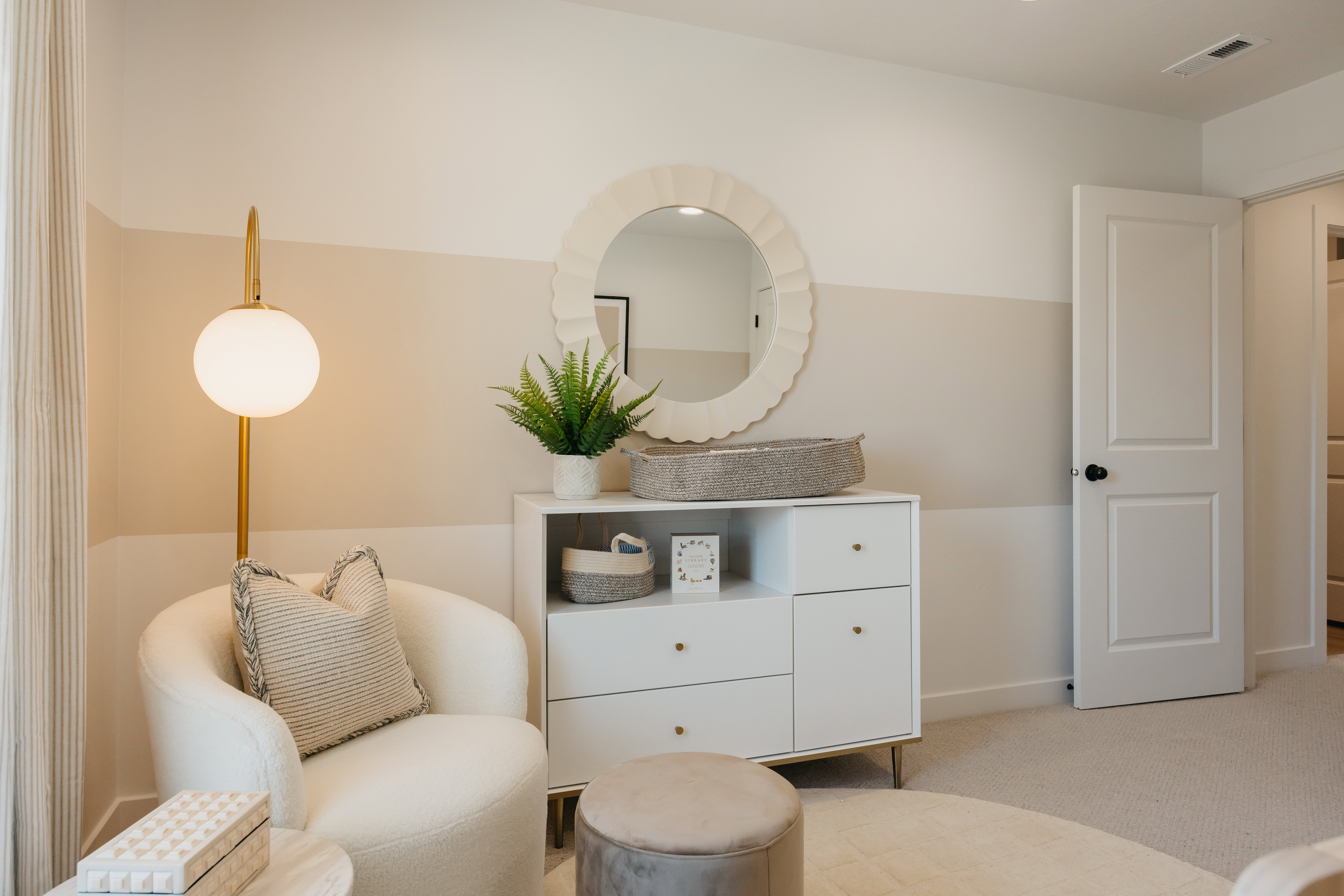 Warmly lit interior space featuring a white dresser with gold hardware, a decorative circular mirror, and a modern floor lamp