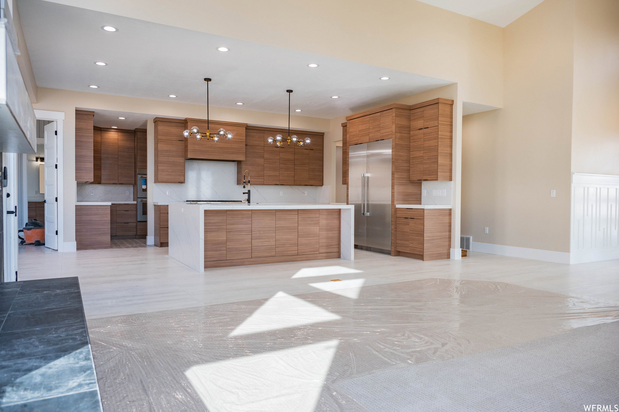 Open-concept kitchen featuring wood-finish cabinetry, a large center island with a white countertop, stainless steel appliances, and two contemporary chandeliers