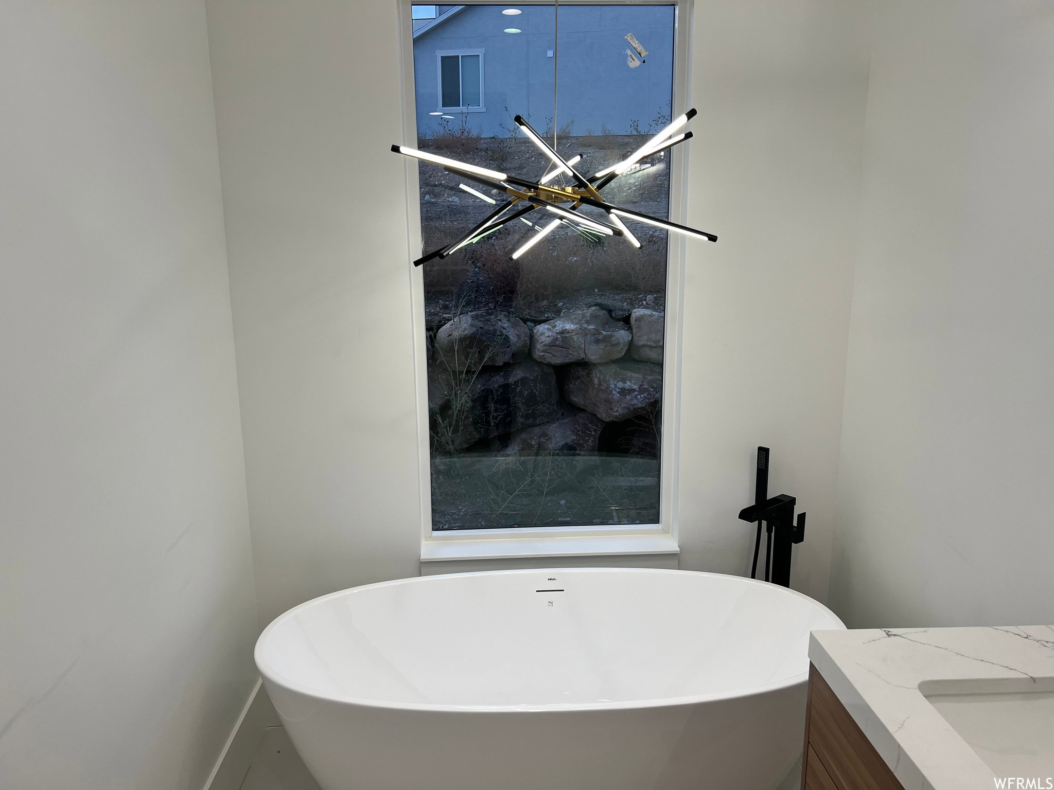 Freestanding soaking tub with a floor-mounted matte black faucet, set against a window and under a contemporary linear chandelier