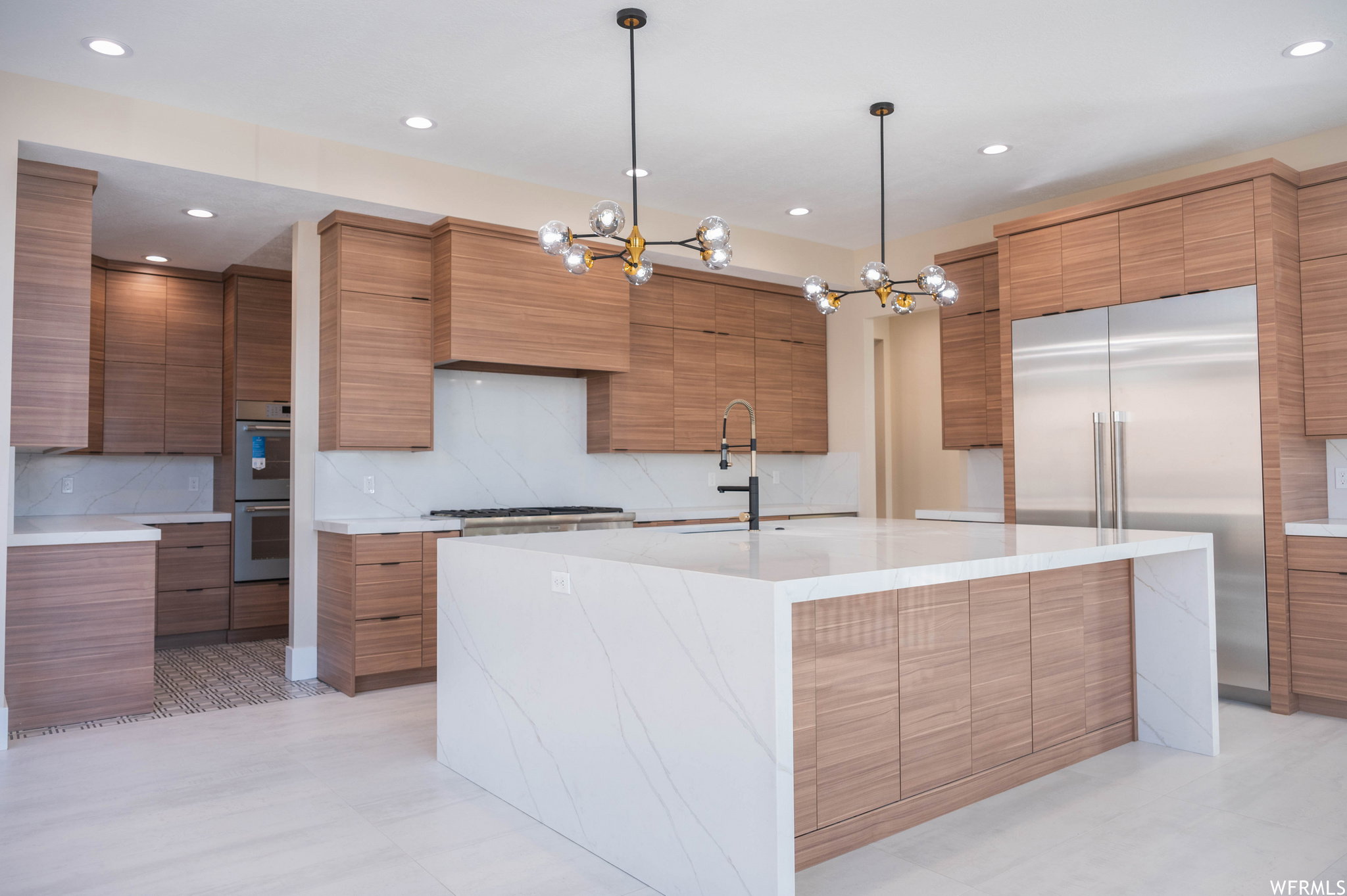 Expansive kitchen featuring a large waterfall island with integrated cabinetry, full-height tile backsplash, stainless steel appliances, and wood-finish cabinetry