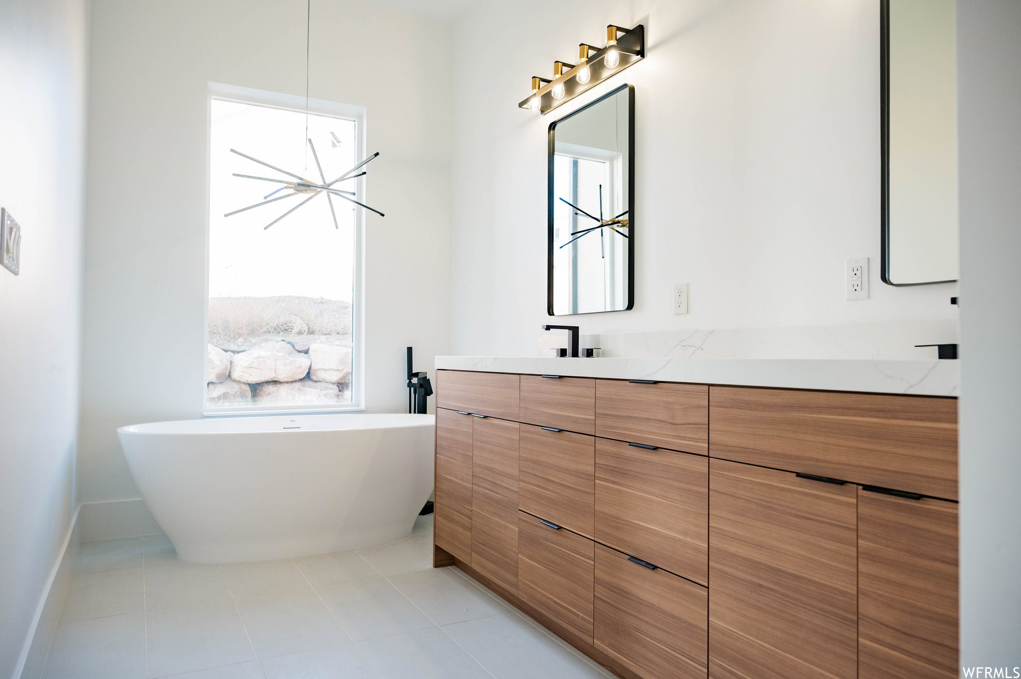 Modern bathroom featuring a freestanding soaking tub, a multi-light linear chandelier, and a dual vanity with wood-finish cabinetry, white countertops, and black framed mirrors