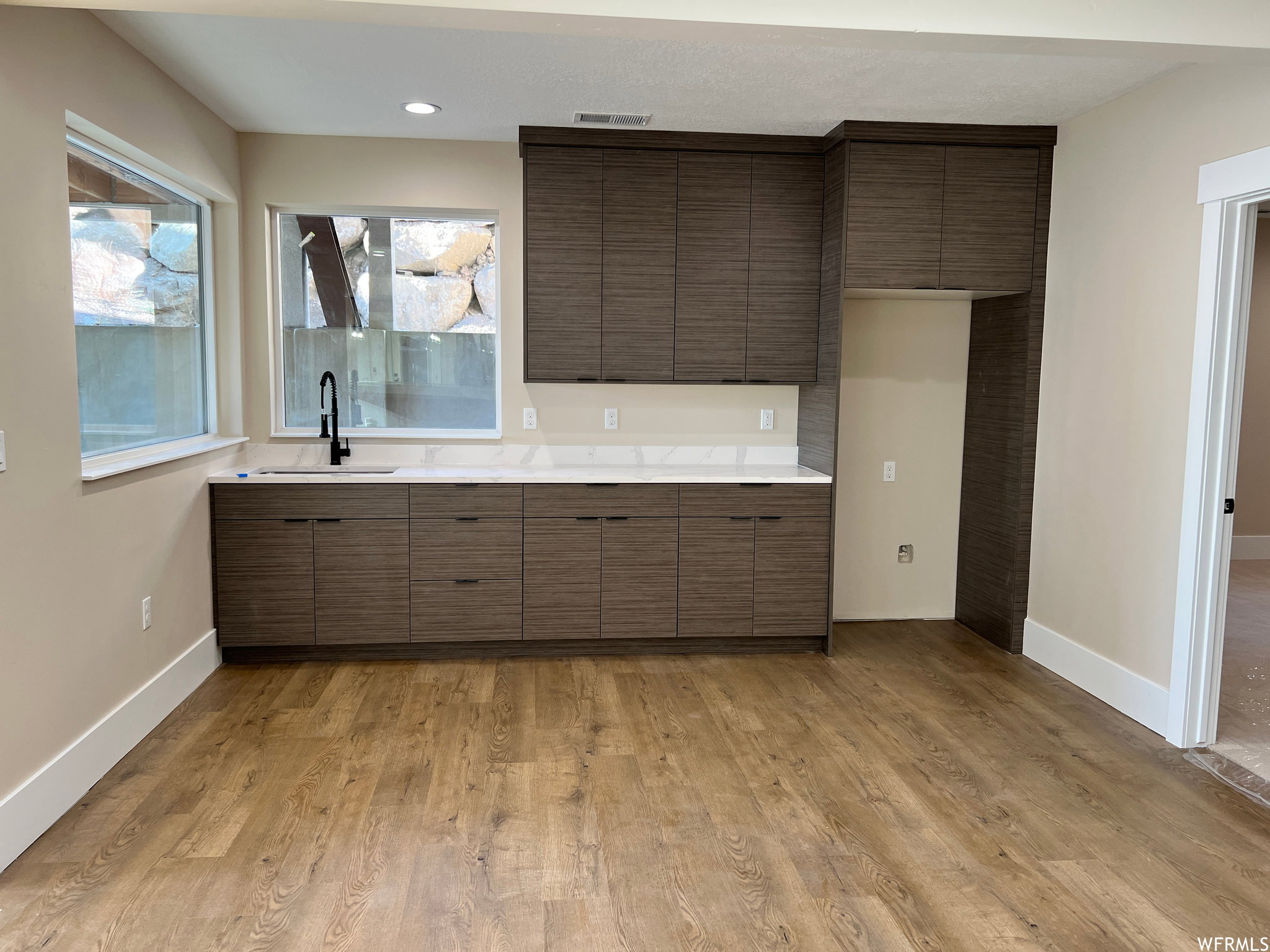 Kitchenette featuring wood-finish flooring, dark wood-finish cabinetry, light-toned countertops, a matte black faucet, and recessed lighting