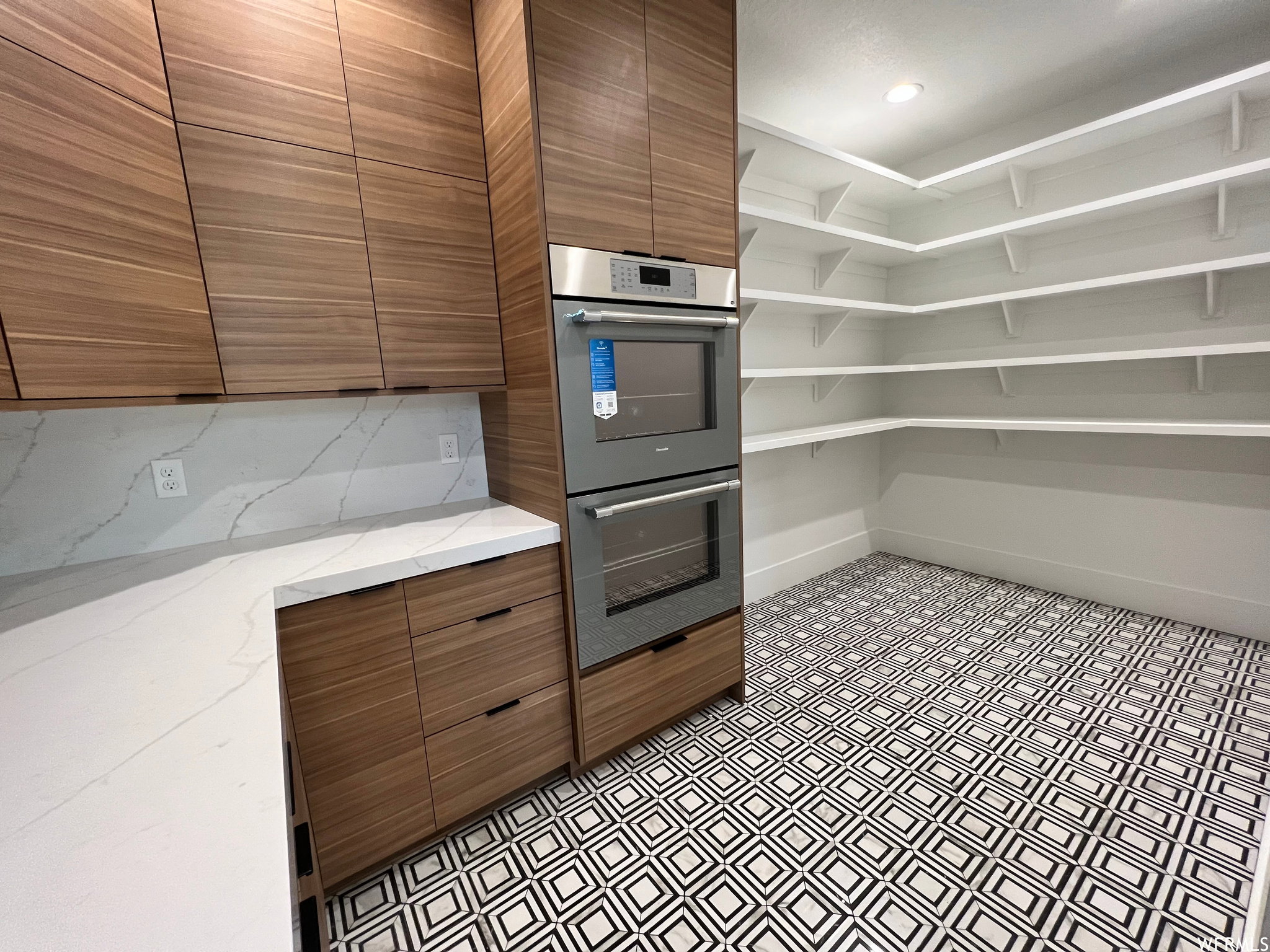 Kitchen pantry featuring extensive built-in shelving, wood-finish cabinetry, a double wall oven, quartz-finish countertops, and patterned floor tiling