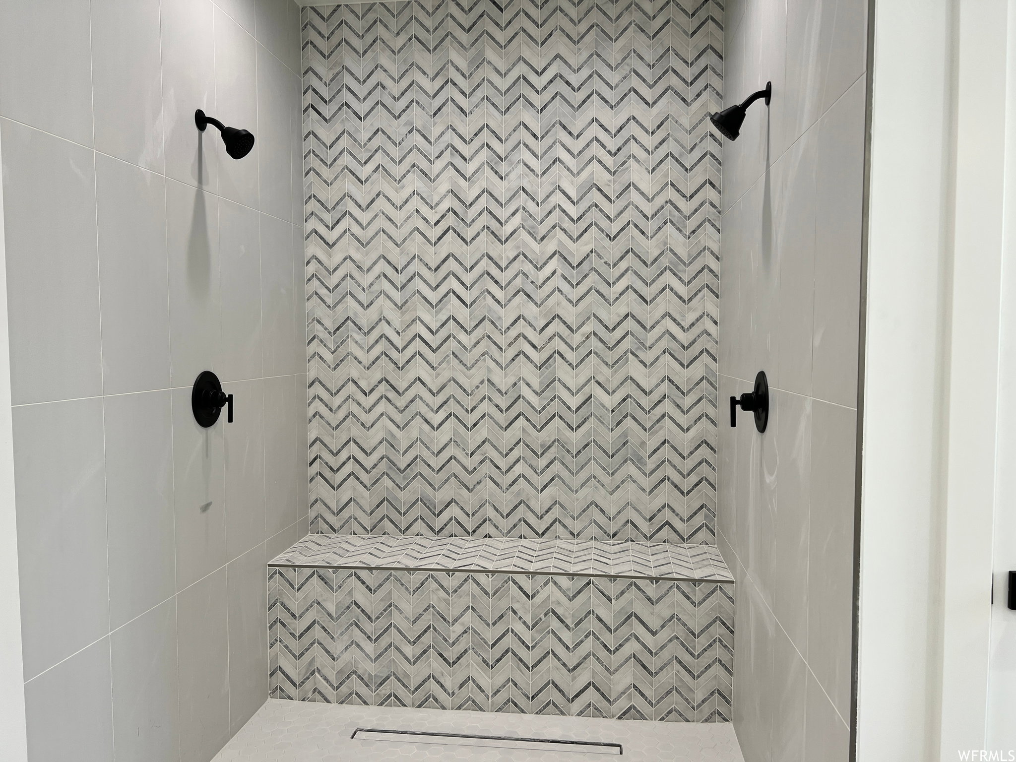 Walk-in shower with chevron-patterned tile accent wall, integrated shower bench, dual showerheads with matte black fixtures, and large format wall tiling