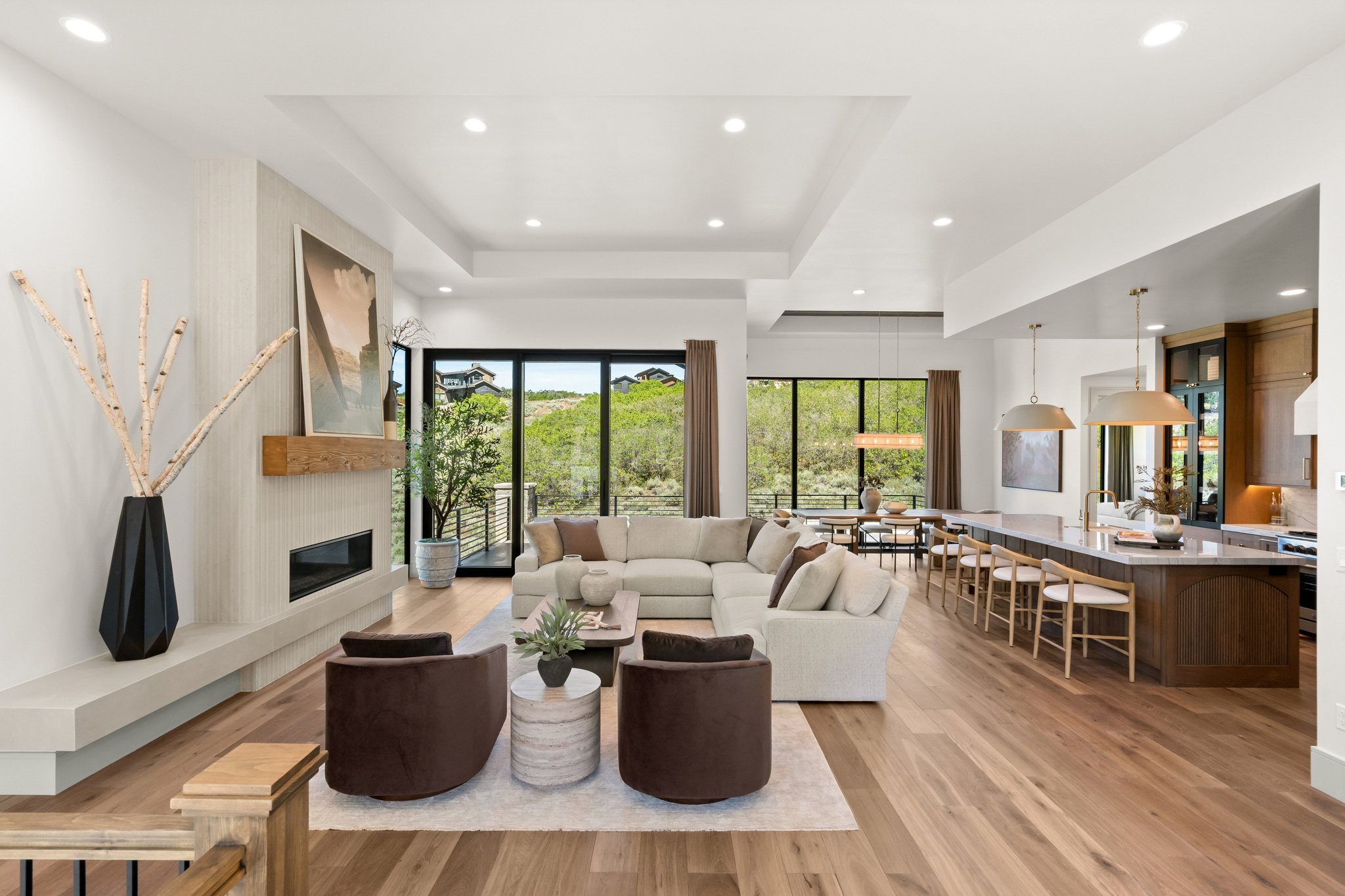 Open-concept living space featuring wood-finish flooring, a linear fireplace with a stone surround and wood mantel, large glass sliding doors, recessed lighting, and a kitchen island with seating