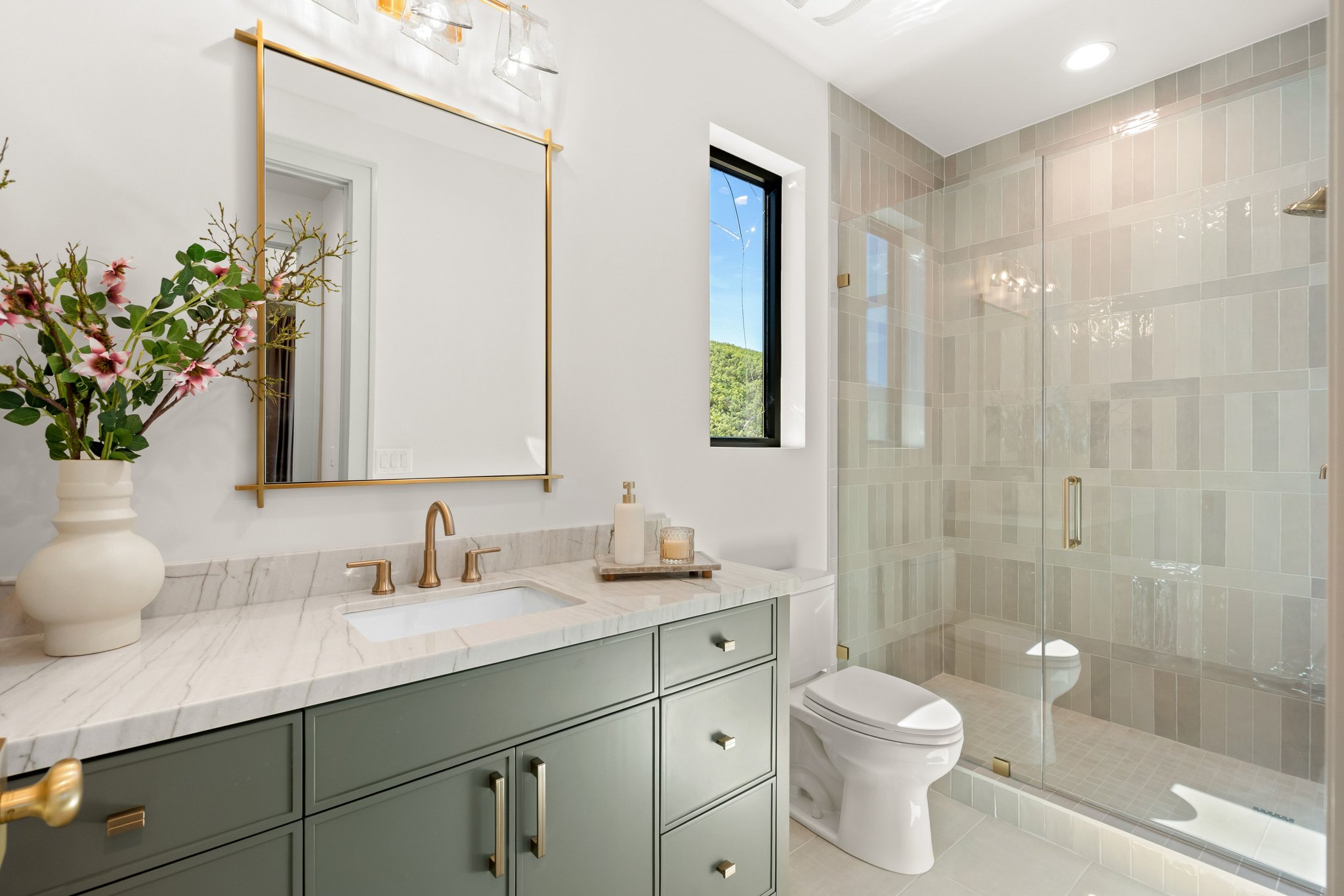 Bathroom featuring a sage green vanity with a white countertop and gold-toned hardware, a frameless glass shower enclosure with vertical tile accents, a rectangular window with black trim, a gold-framed mirror, and a contemporary vanity light fixture