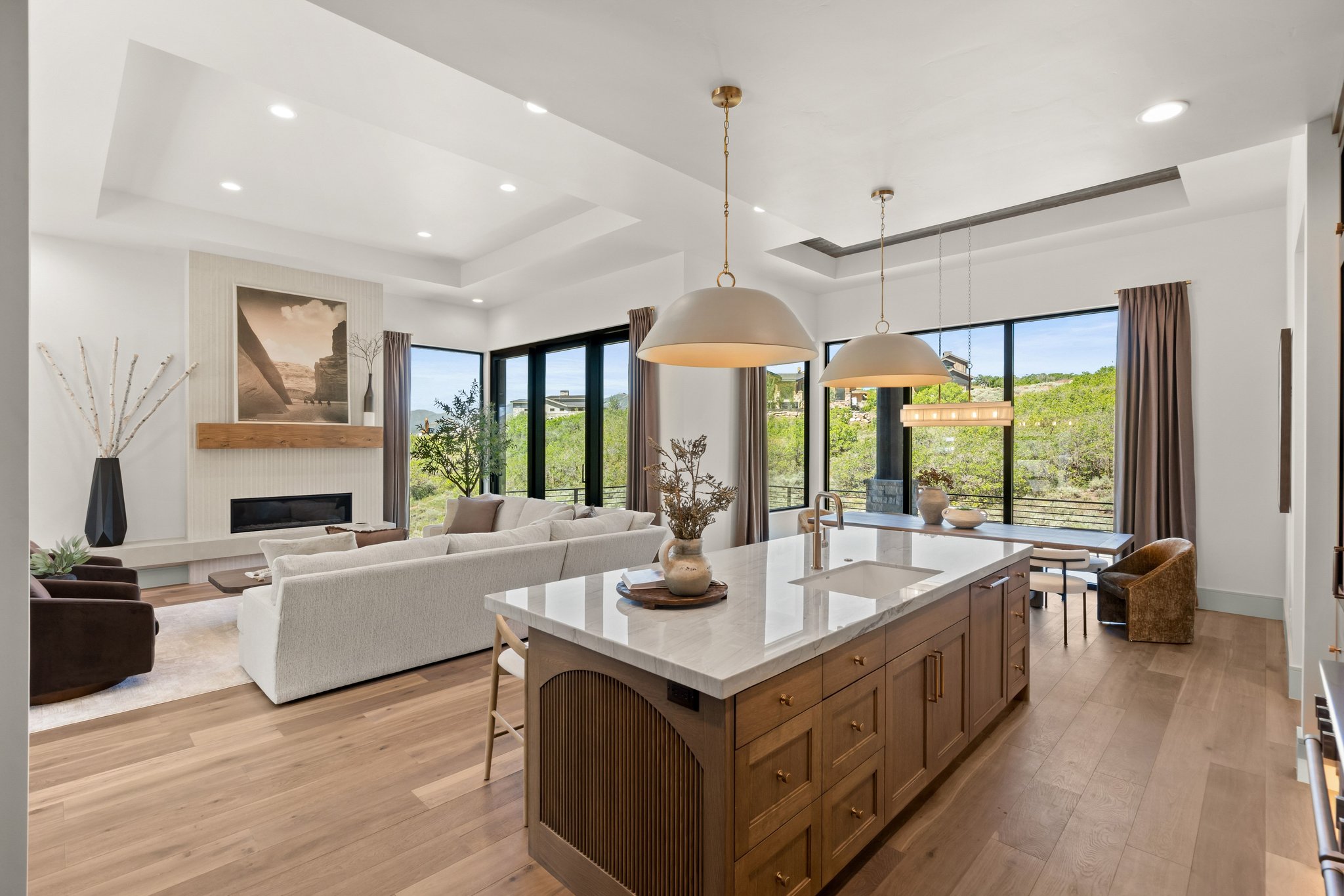 Open-concept interior featuring wood-finish flooring, a kitchen island with an undermount sink, a recessed fireplace with a wood mantle, and expansive windows