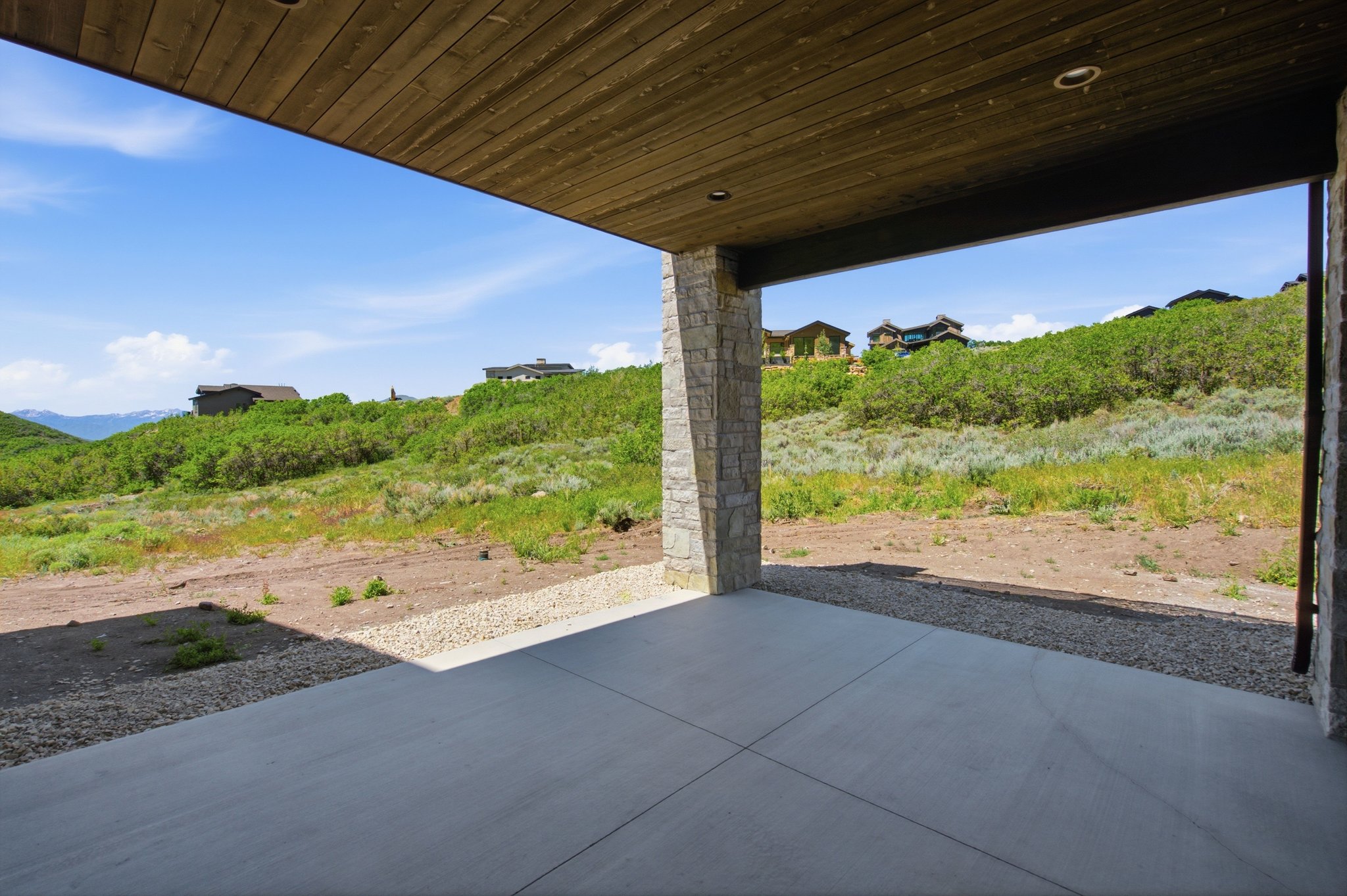 Covered patio featuring a concrete surface, stone-clad columns, and a wood-finish ceiling with recessed lighting, overlooking a natural hillside landscape