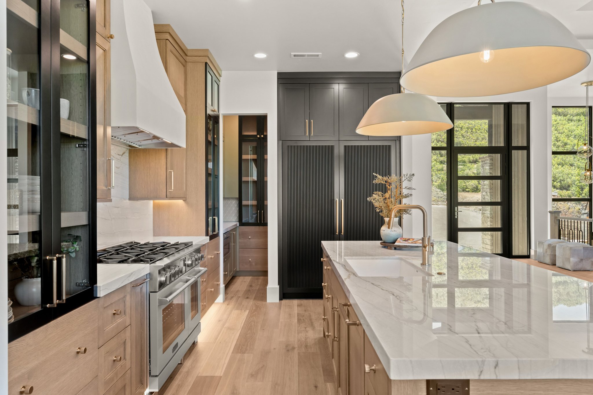 Gourmet kitchen featuring a large central island with an undermount sink and brass faucet, light wood-finish cabinetry with brass hardware, integrated stainless steel appliances, a white range hood, and black-framed glass paneled doors