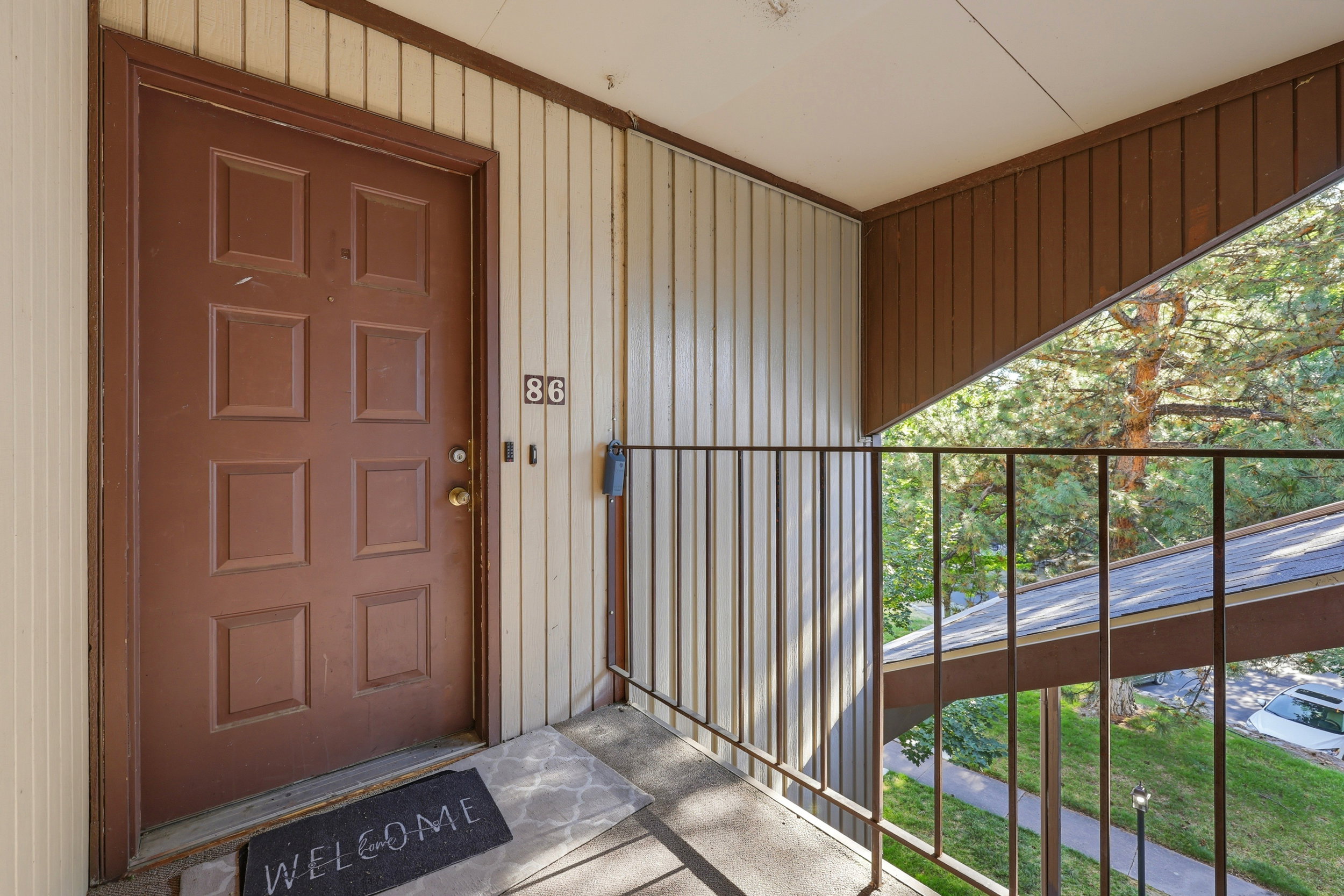Entry door with multiple panels, exterior wall with vertical siding, outdoor balcony with metal railing, and tree-lined backdrop