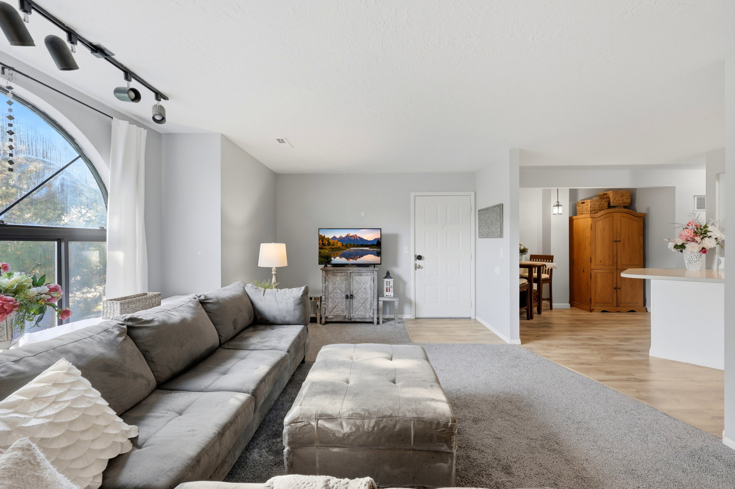 Spacious living area featuring a large arched window, track lighting, light gray wall paint, and mixed flooring with carpet and wood-finish