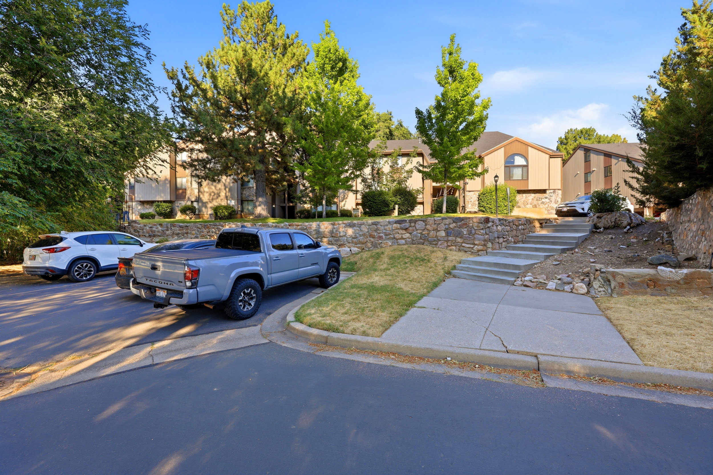 Property exterior featuring tan siding, a stone retaining wall, mature trees, and concrete steps